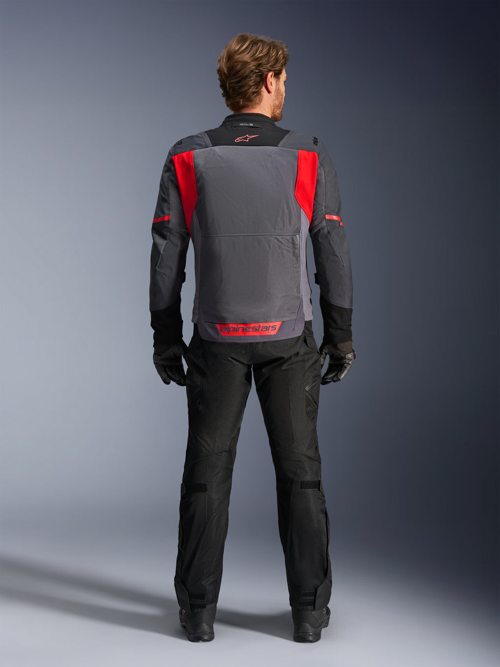 Giacca Alpinestars ST-2 Air, Deep Gray, Dark Gray e Bright Red, vista posteriore indossata da un pilota con pantaloni e guanti neri, dotata di ampi pannelli di ventilazione in rete e logo Alpinestars rosso sulla parte inferiore della schiena, design moderno per touring estivo