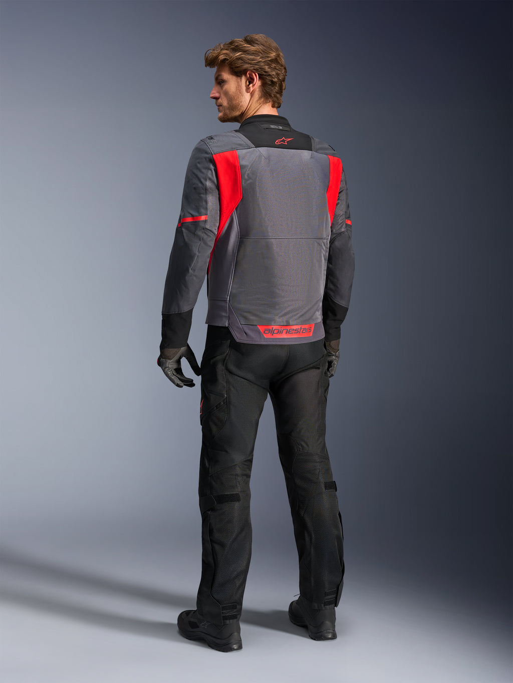 Pilota visto da dietro che indossa la giacca Alpinestars St-2 Air in Deep Gray, Dark Gray e Bright Red, che mette in risalto l'ampio pannello di ventilazione in rete sulla schiena e il logo rosso, abbinata a pantaloni da moto neri e guanti grigi.