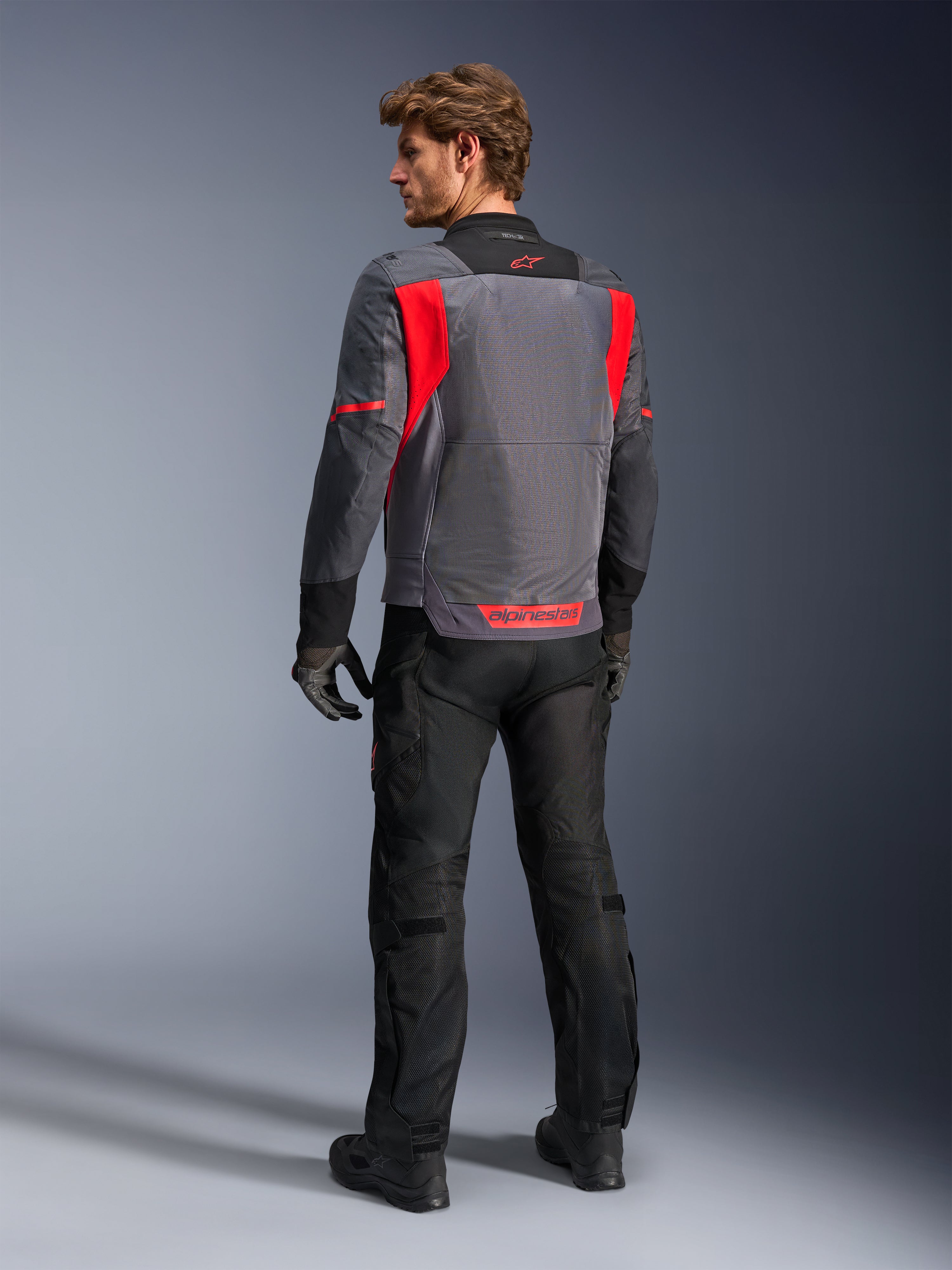 Pilota visto da dietro che indossa la giacca Alpinestars St-2 Air in Deep Gray, Dark Gray e Bright Red, che mette in risalto l'ampio pannello di ventilazione in rete sulla schiena e il logo rosso, abbinata a pantaloni da moto neri e guanti grigi.