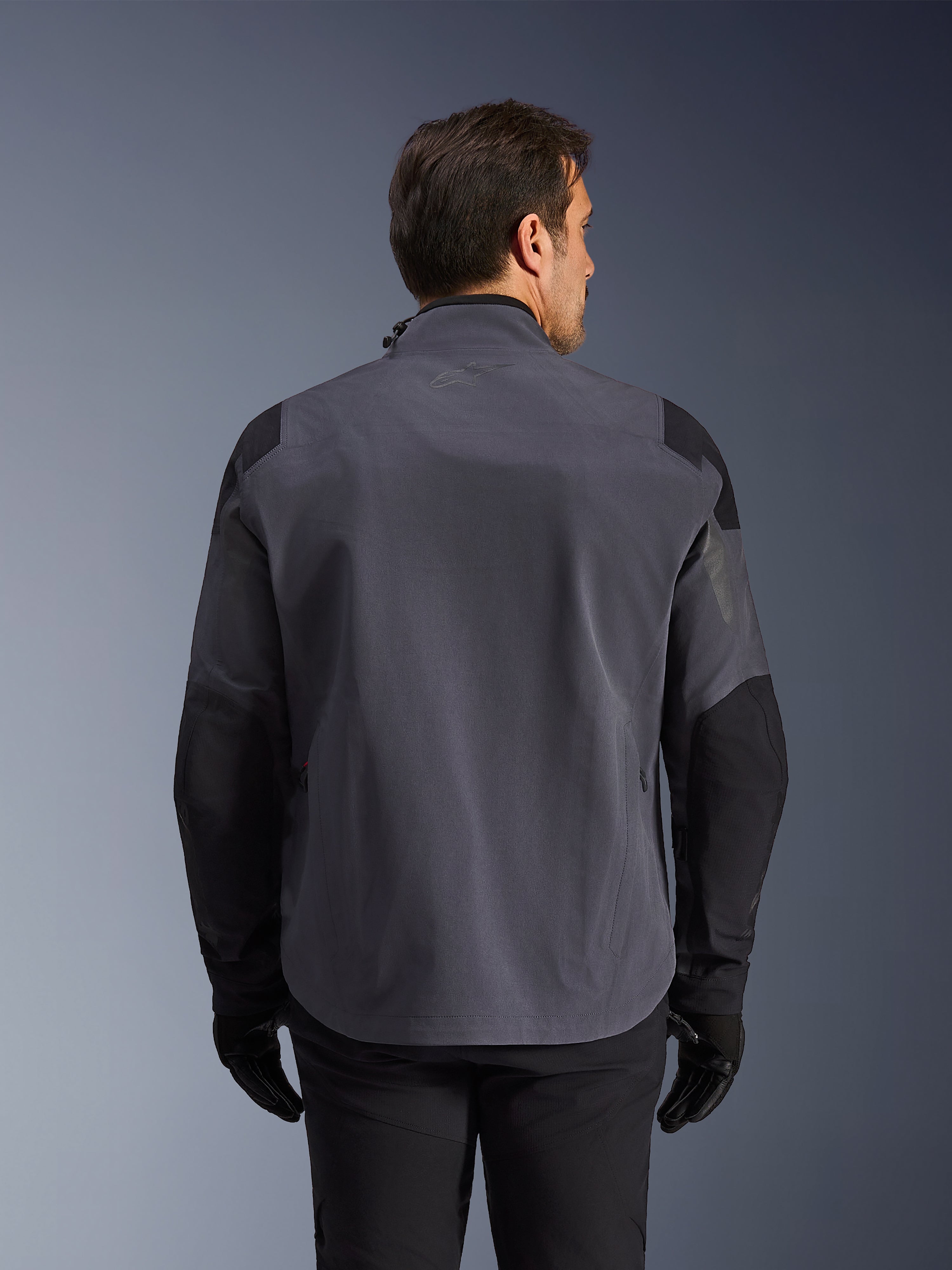 Alpinestars Maxdura Drystar®XF Laminated Armored Shell, giacca tecnica adventure, grigio scuro e nero, vista posteriore con pannelli rinforzati su spalle e gomiti, logo discreto sulla parte superiore della schiena, indossata con guanti e pantaloni neri, progettata per la protezione in ogni condizione atmosferica