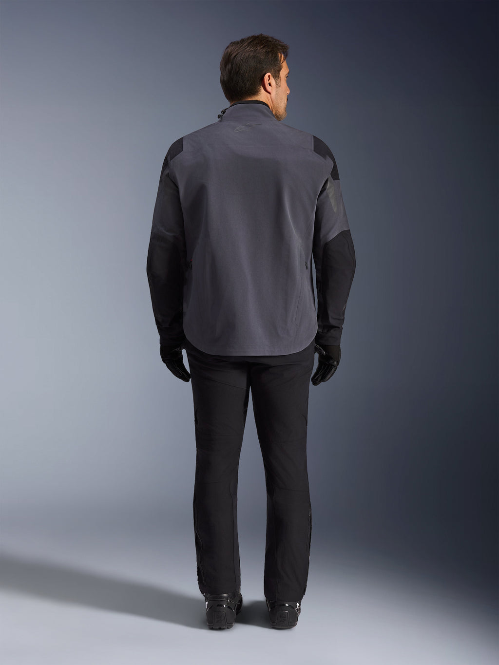 Alpinestars Maxdura Drystar®XF Laminated Armored Shell, giacca da moto, grigio scuro e nero, vista posteriore di un pilota con equipaggiamento adventure completo, guscio laminato tecnico con pannelli neri rinforzati su spalle e gomiti, abbinata a pantaloni e guanti neri.