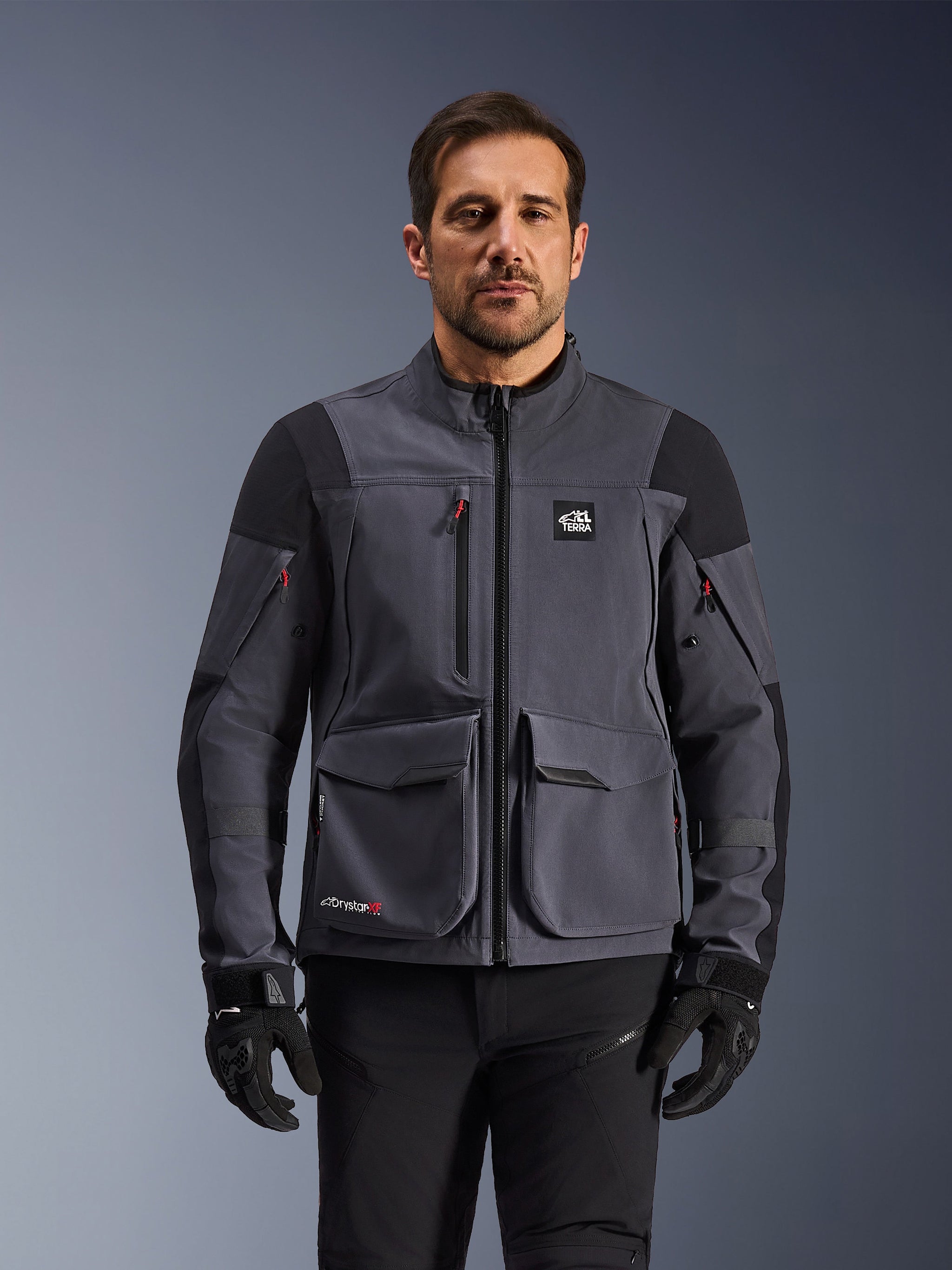 Alpinestars Maxdura Drystar®XF Laminated Armored Shell, giacca da moto adventure in grigio scuro e nero, vista frontale su modello, con ampie tasche cargo, logo 'All Terra' sul petto e cerniere di ventilazione, abbinata a guanti e pantaloni da guida neri.