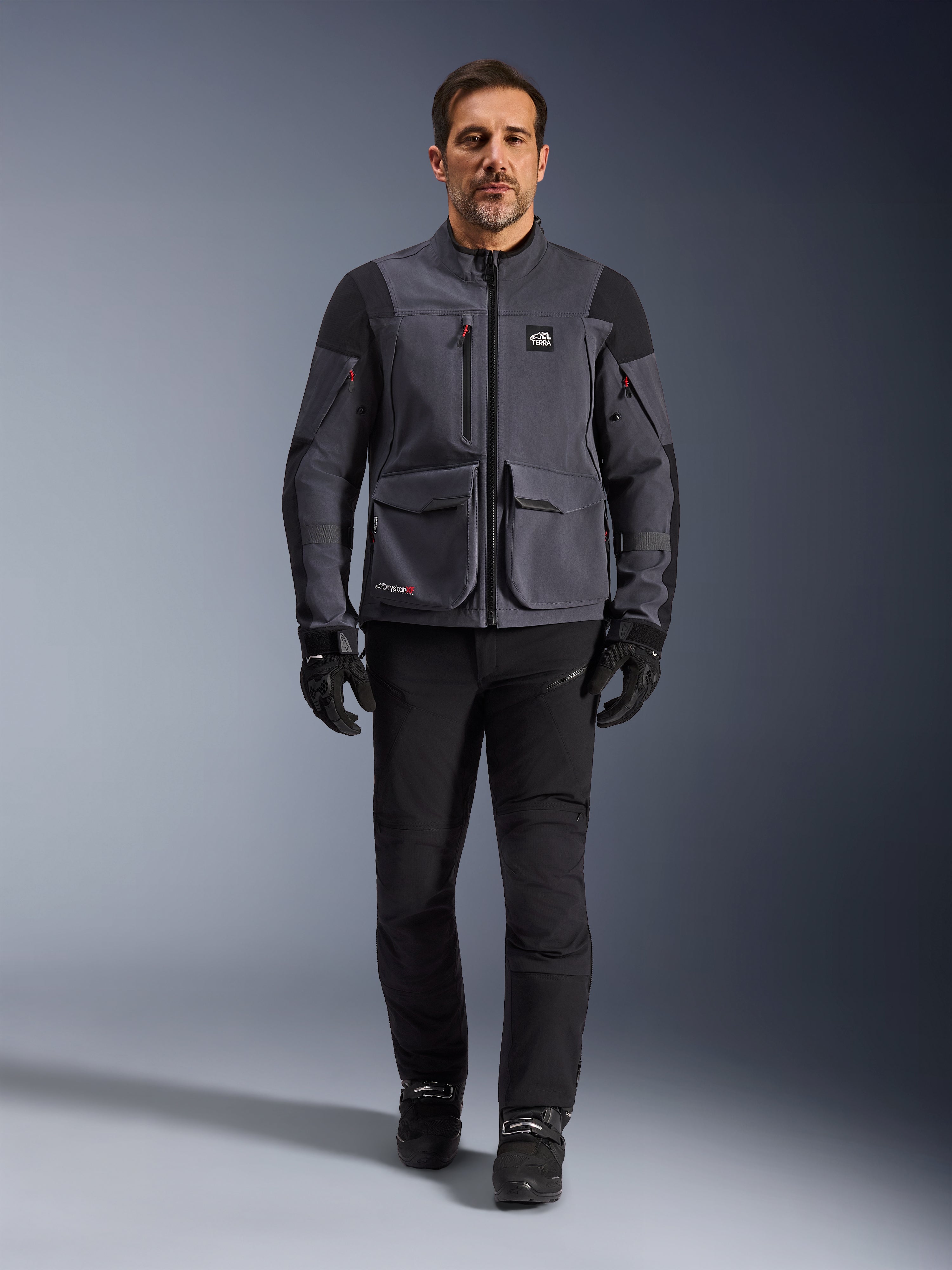 Maxdura Drystar®XF Laminated Armored Shell, giacca da moto, Deep Gray Black, indossata da un modello con pantaloni e guanti neri, con ampie tasche cargo, dettagli delle cerniere rossi e marchio All Terra, abbigliamento tecnico da moto adventure