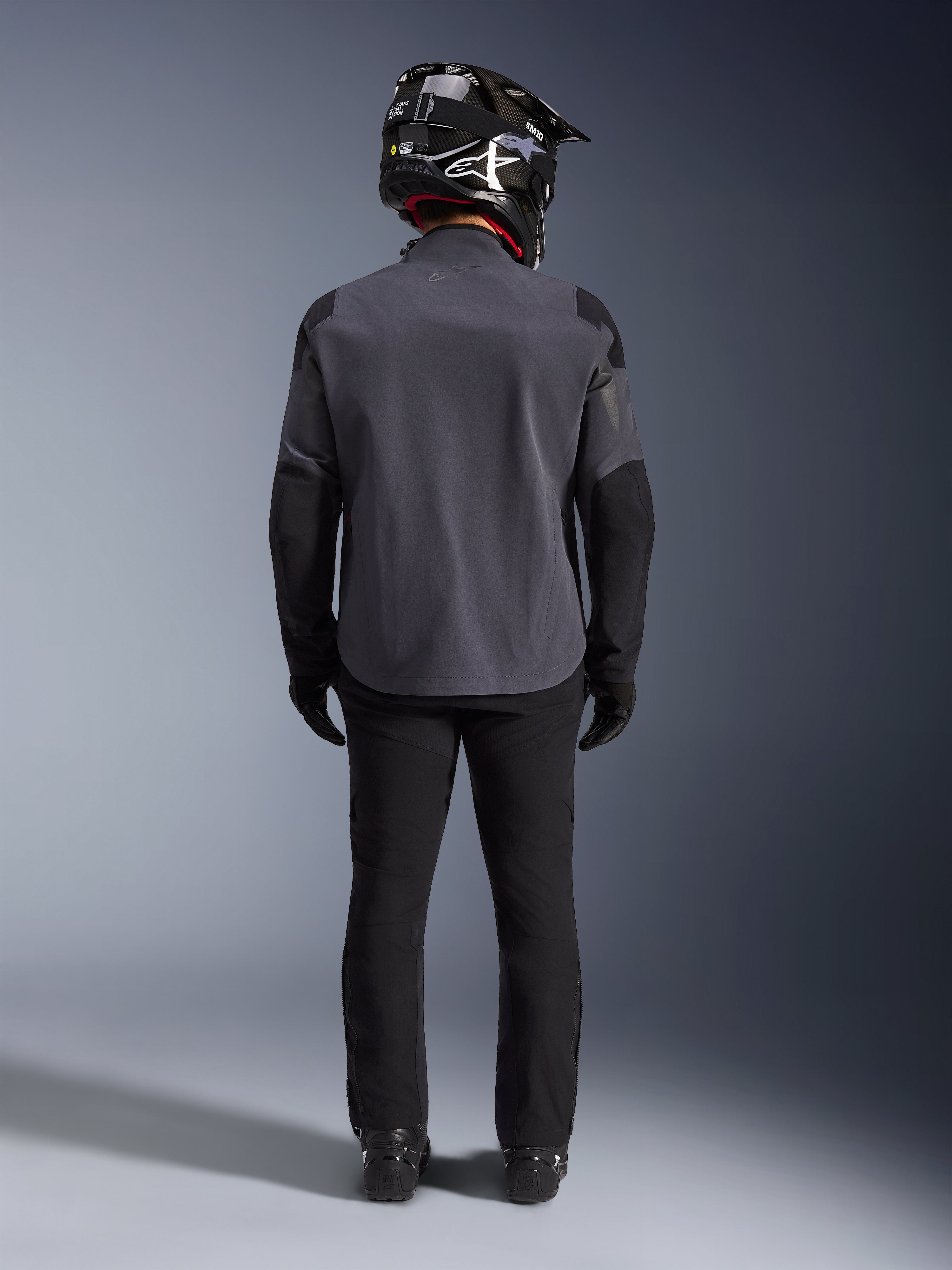 Alpinestars Maxdura Drystar®XF Laminated Armored Shell, Deep Gray Black, vista posteriore a figura intera, giacca tecnica adventure con pannelli rinforzati, indossata con pantaloni da guida neri, guanti e casco off-road bianco e nero, completo coordinato dual-sport