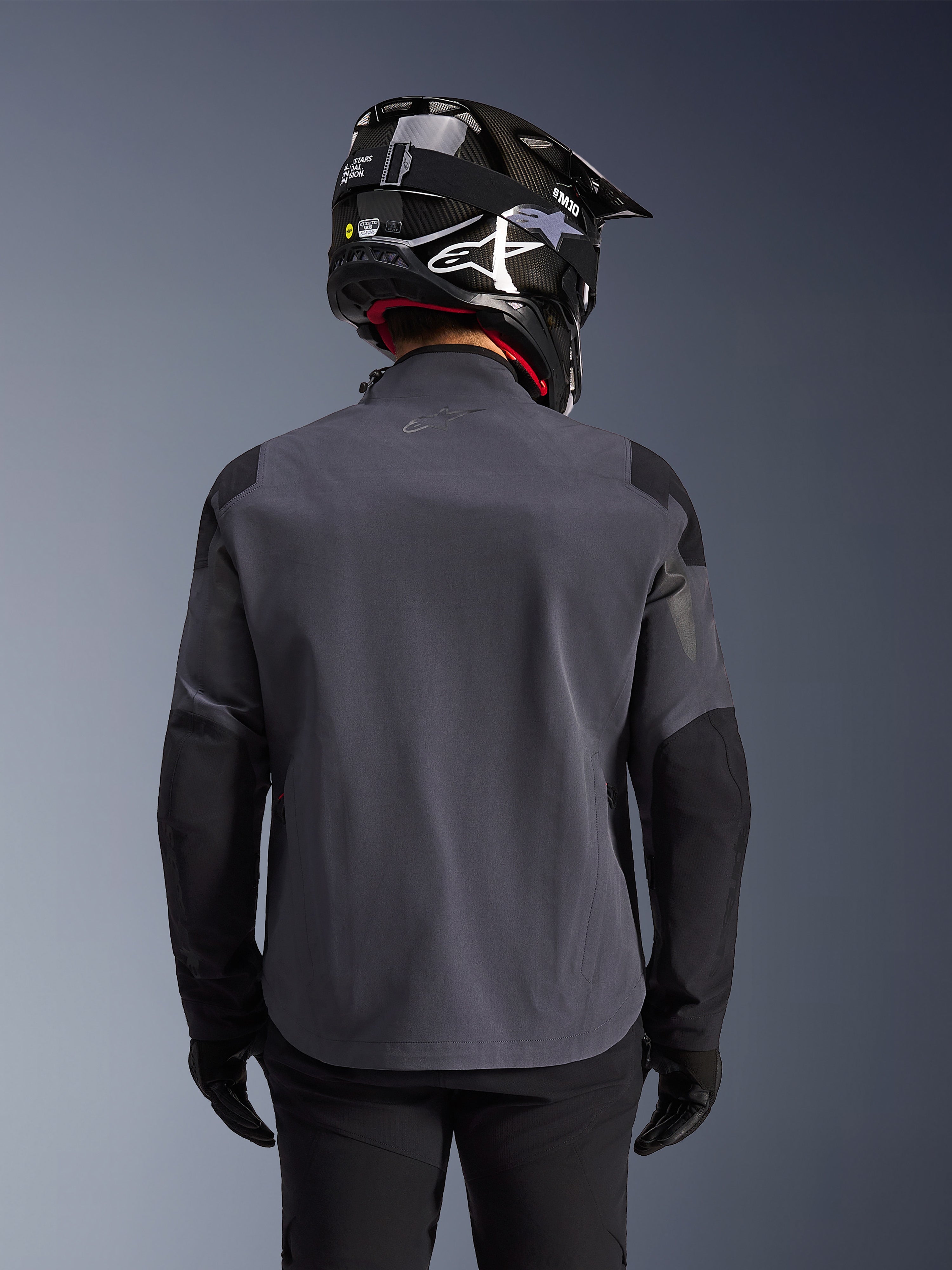 Maxdura Drystar®XF Laminated Armored Shell, giacca da moto adventure, Deep Gray Black, vista posteriore che mostra la costruzione in tessuto tecnico e i pannelli rinforzati, indossata con casco in fibra di carbonio nero, pantaloni da guida neri e guanti