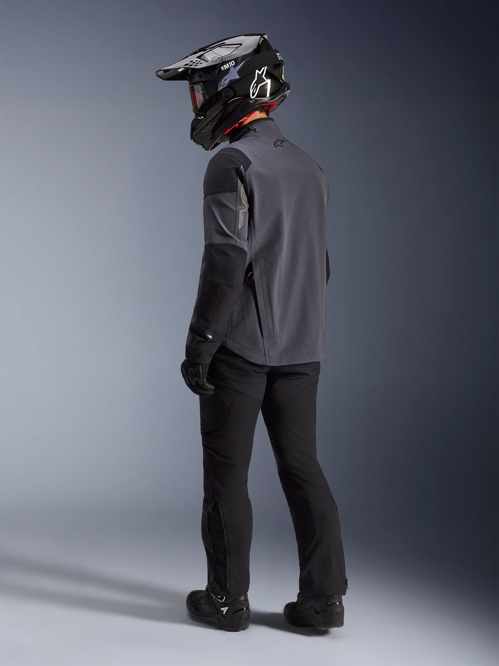 Pilota adventure che indossa la giacca Maxdura Drystar®XF Laminated Armored Shell in Deep Gray Black, vista posteriore. Equipaggiamento completo che include pantaloni da moto neri, un casco da motocross nero con marchio Alpinestars e stivali off-road neri. Kit tecnico per ogni condizione atmosferica per prestazioni dual-sport.