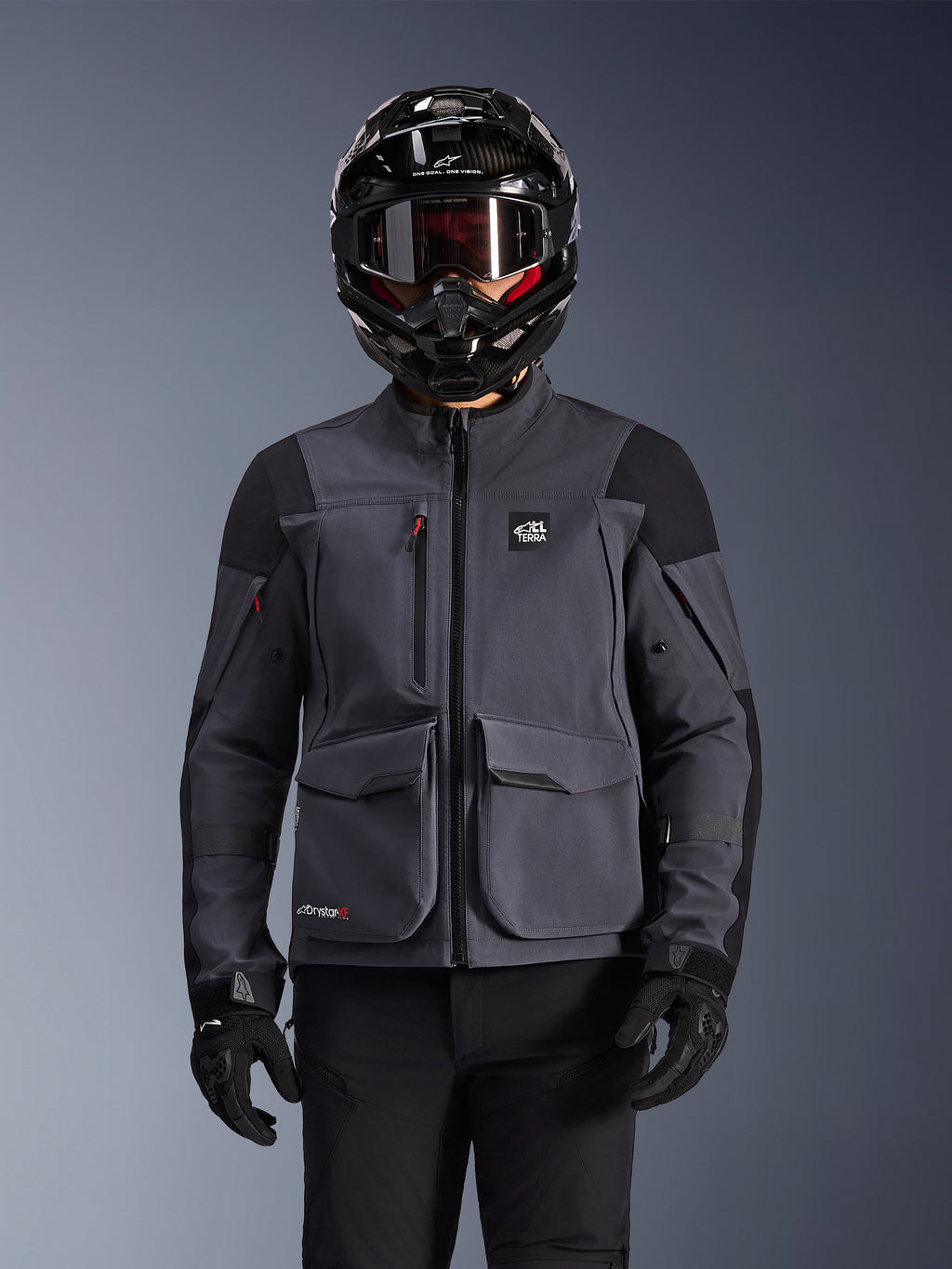 Alpinestars Maxdura Drystar®XF Laminated Armored Shell, giacca da moto, grigio scuro e nero, indossata da un pilota con casco nero, maschera e guanti, dotata di ampie tasche cargo tecniche e marchio ALL TERRA, abbigliamento adventure touring