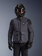 Alpinestars Maxdura Drystar®XF Laminated Armored Shell, giacca da moto, grigio scuro e nero, indossata da un pilota con casco nero, maschera e guanti, dotata di ampie tasche cargo tecniche e marchio ALL TERRA, abbigliamento adventure touring