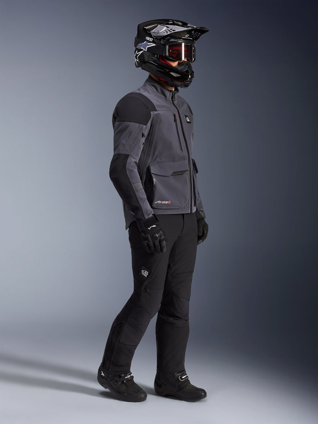 Motociclista che indossa la giacca Alpinestars Maxdura Drystar®XF Laminated Armored Shell in grigio scuro e nero, abbinata a pantaloni da avventura neri, casco nero con occhiali, guanti e stivali neri, scatto a figura intera che evidenzia l'equipaggiamento tecnico dual-sport.