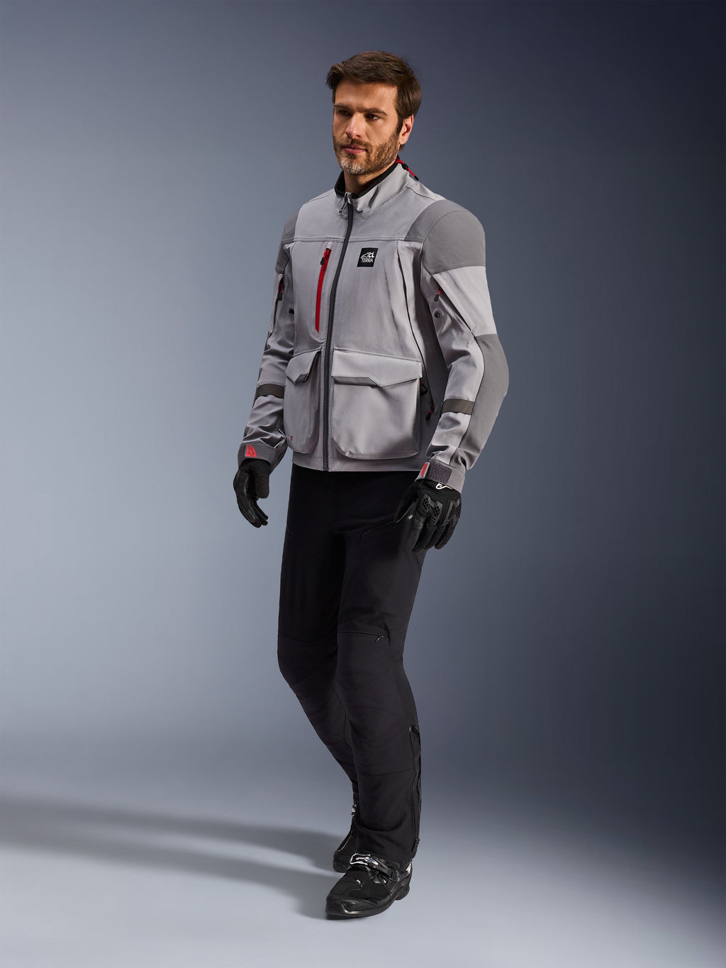 Alpinestars Maxdura Drystar®XF Laminated Armored Shell, giacca da moto adventure, grigio medio e grigio scuro, indossata da un modello con pantaloni da moto e guanti neri, con dettagli delle cerniere rossi e tasche cargo, abbigliamento tecnico professionale per adventure touring