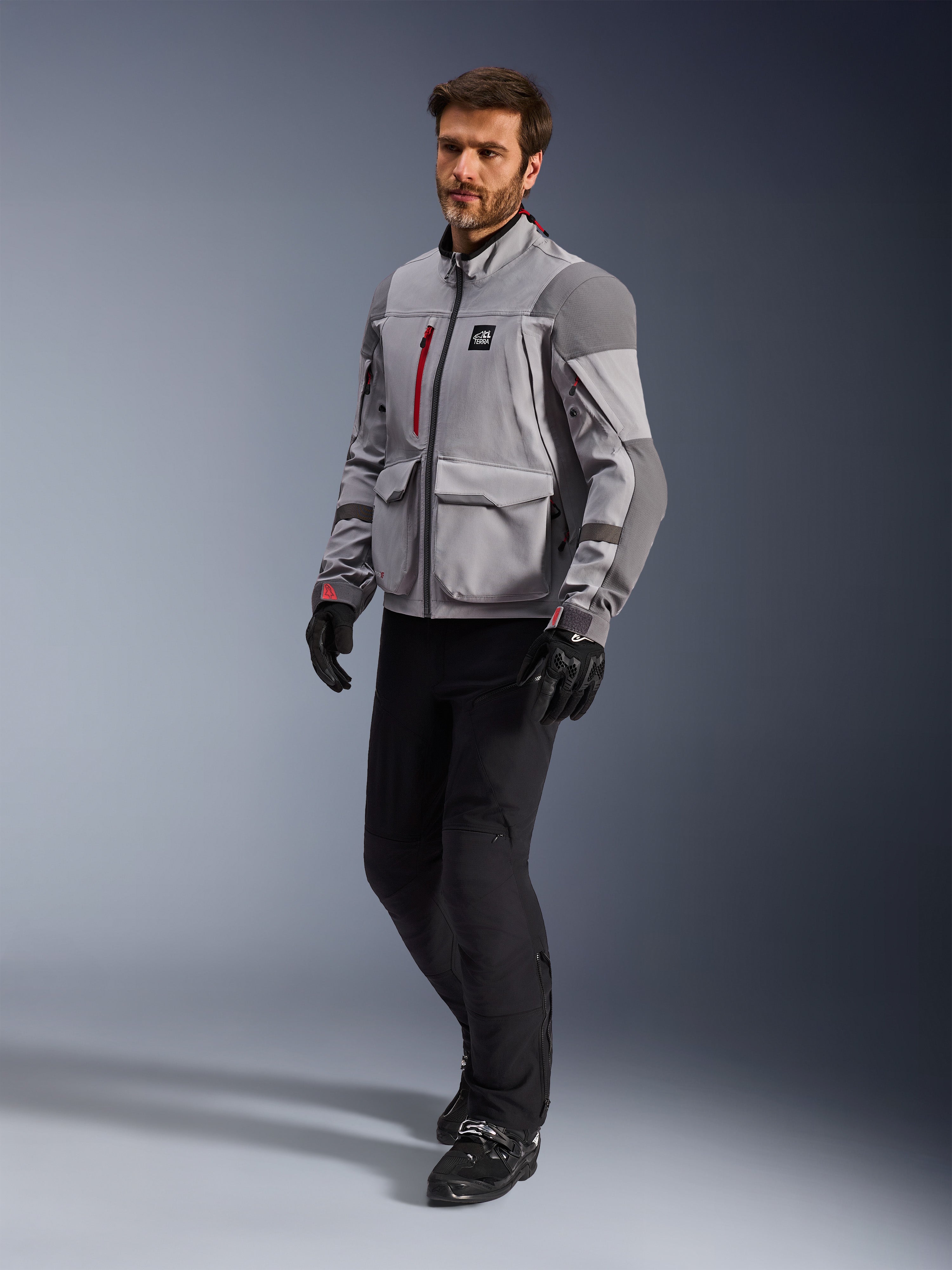 Alpinestars Maxdura Drystar®XF Laminated Armored Shell, giacca da moto adventure, grigio medio e grigio scuro, indossata da un modello con pantaloni da moto e guanti neri, con dettagli delle cerniere rossi e tasche cargo, abbigliamento tecnico professionale per adventure touring