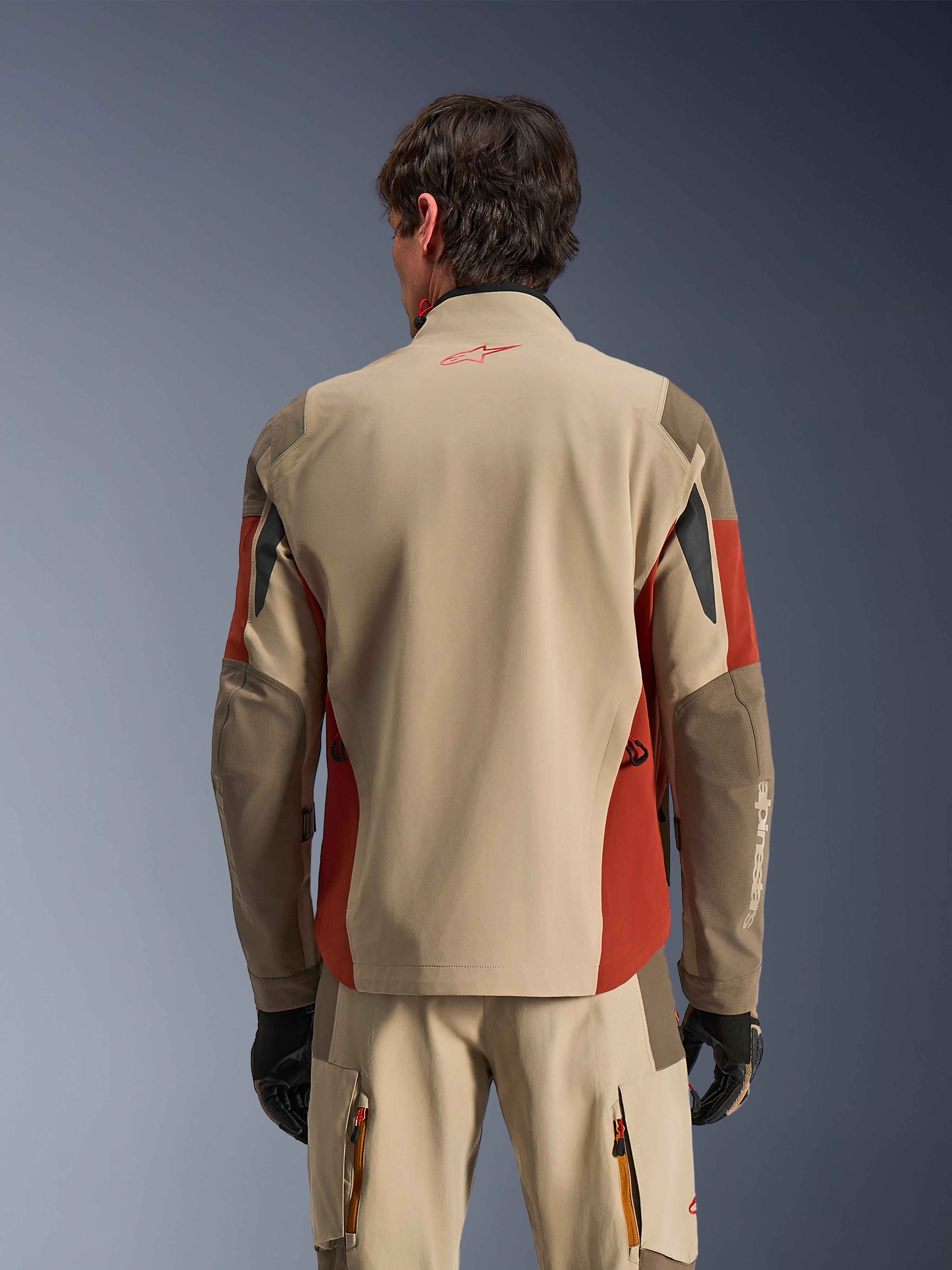 Uomo che indossa Alpinestars Maxdura Drystar®XF Laminated Armored Shell visto da dietro, giacca adventure color Aluminum Walnut e Rust Brown, pantaloni tecnici coordinati e guanti neri, costruzione in tessuto laminato a 3 strati con pannelli rinforzati, completo coordinato per touring motociclistico