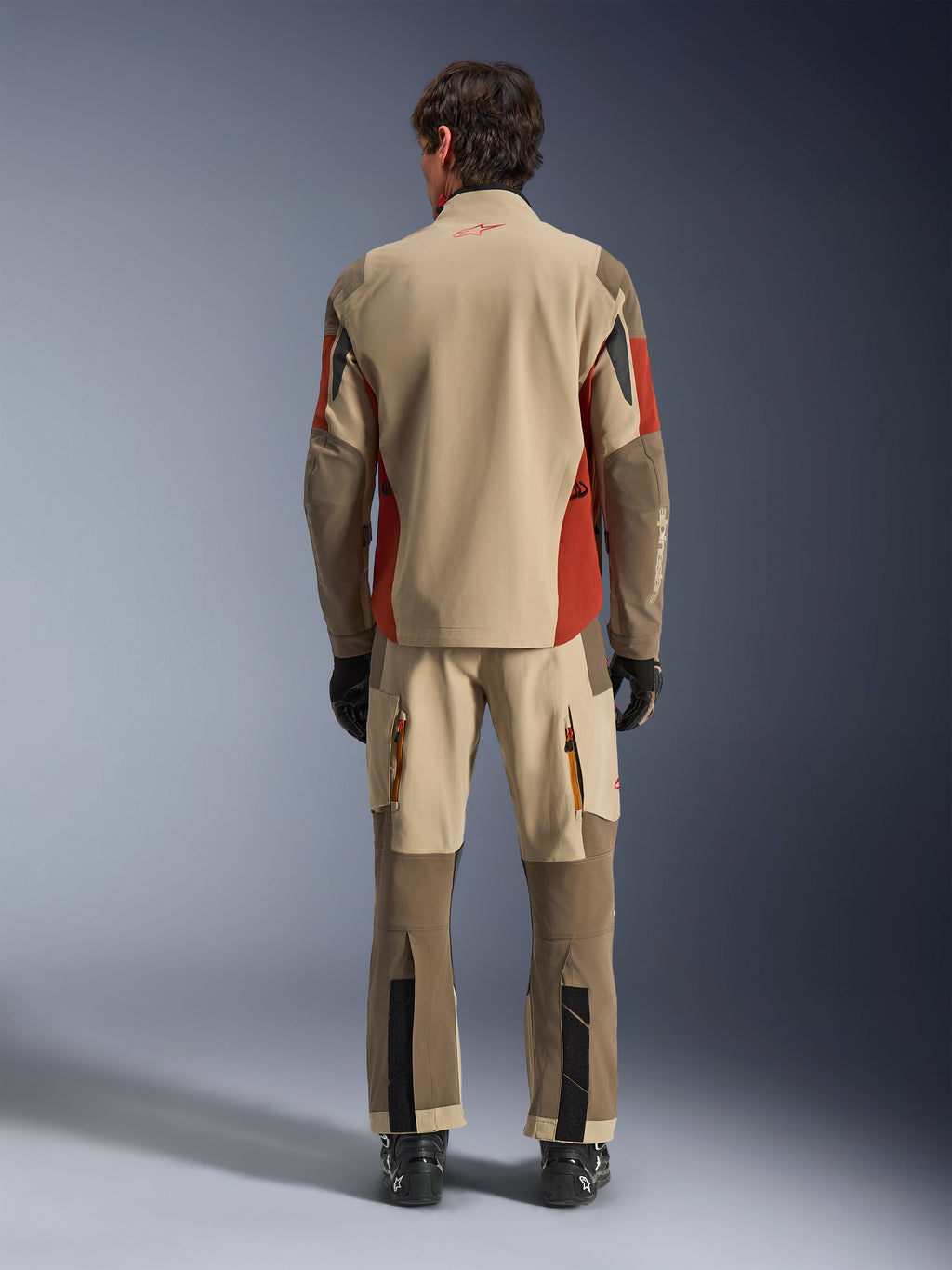 Vista posteriore di un pilota che indossa la giacca Alpinestars Maxdura Drystar®XF Laminated Armored Shell e pantaloni adventure coordinati in Aluminum Walnut Rust Brown, tessuto marrone e color sabbia con accenti arancioni, abbinati a stivali e guanti da moto neri, equipaggiamento professionale da adventure touring.