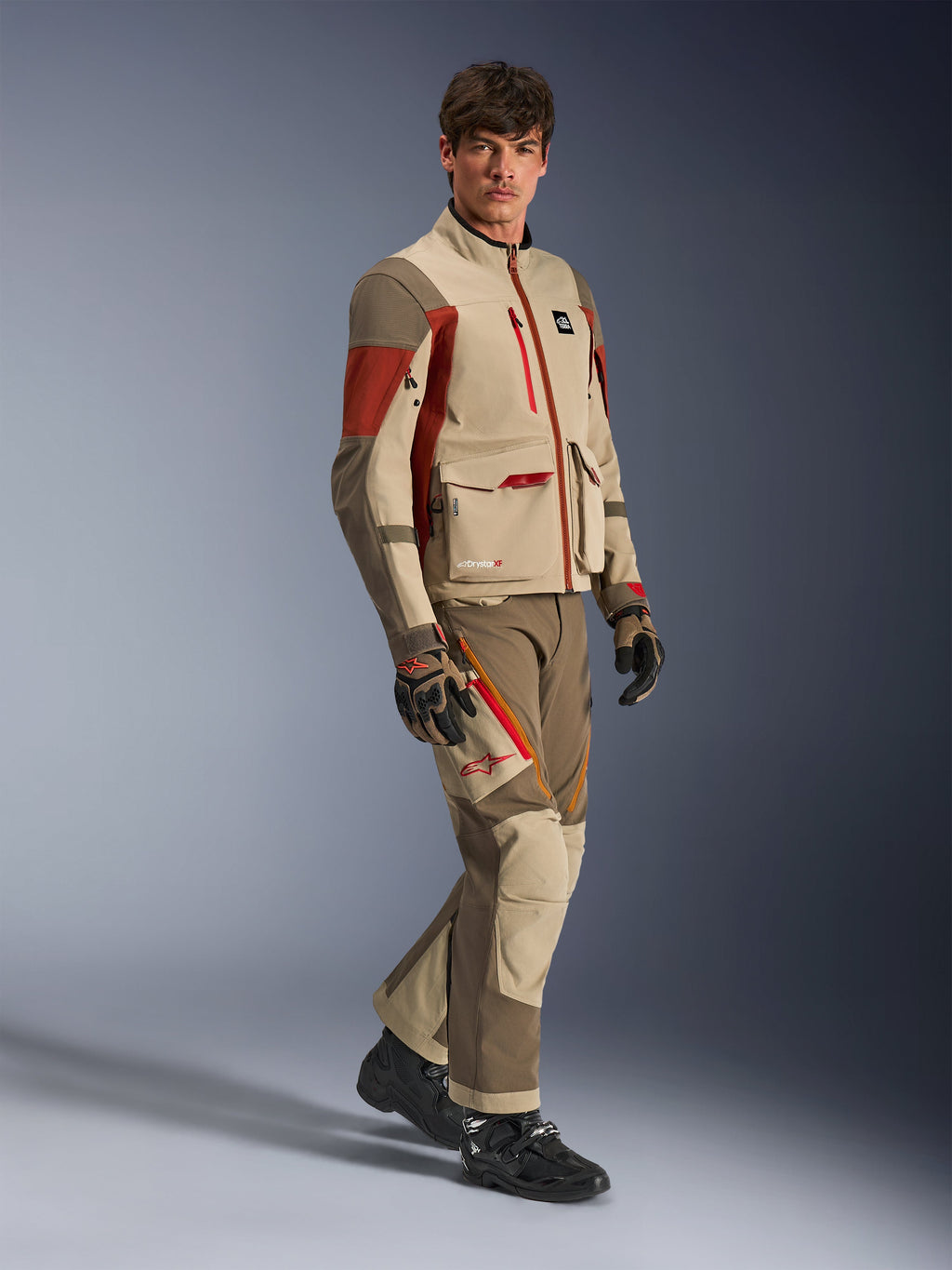 Modello che indossa Alpinestars Maxdura Drystar®XF Laminated Armored Shell, giacca da adventure in colore Aluminum Walnut Rust Brown con pantaloni coordinati, guanti neri e marroni e stivali neri, caratterizzata da un design tecnico multistrato con tasche cargo e dettagli di cerniere rosse, outfit coordinato per adventure touring