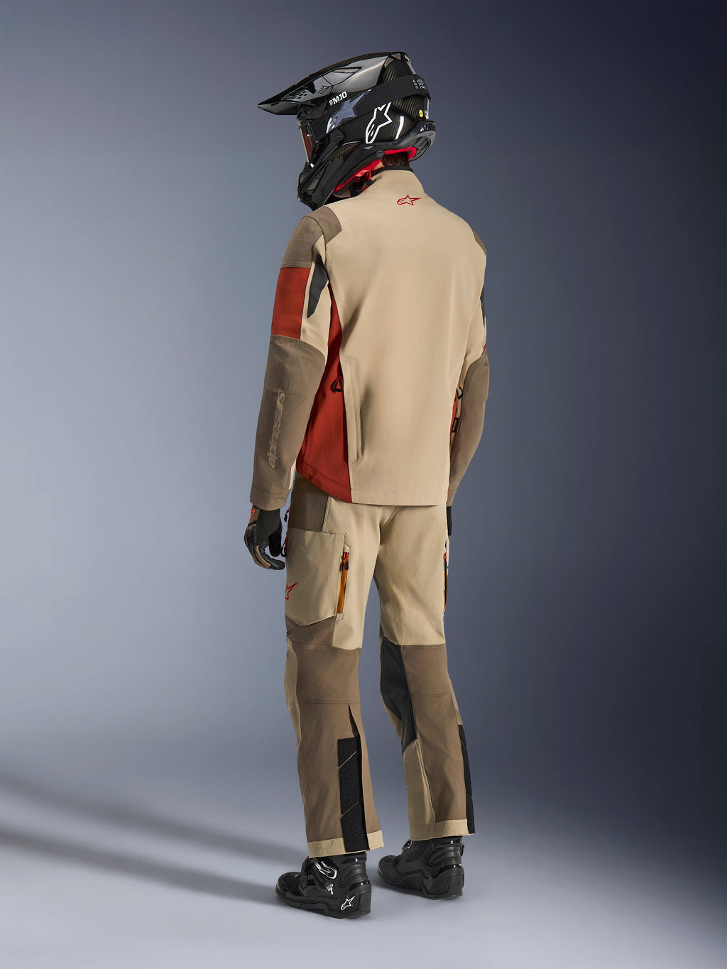 Pilota adventure visto di spalle che indossa la giacca Alpinestars Maxdura Drystar®XF Laminated Armored Shell in Aluminum Walnut Rust Brown, abbinata a pantaloni da moto coordinati, casco off-road nero e stivali neri, outfit tecnico da touring completo.