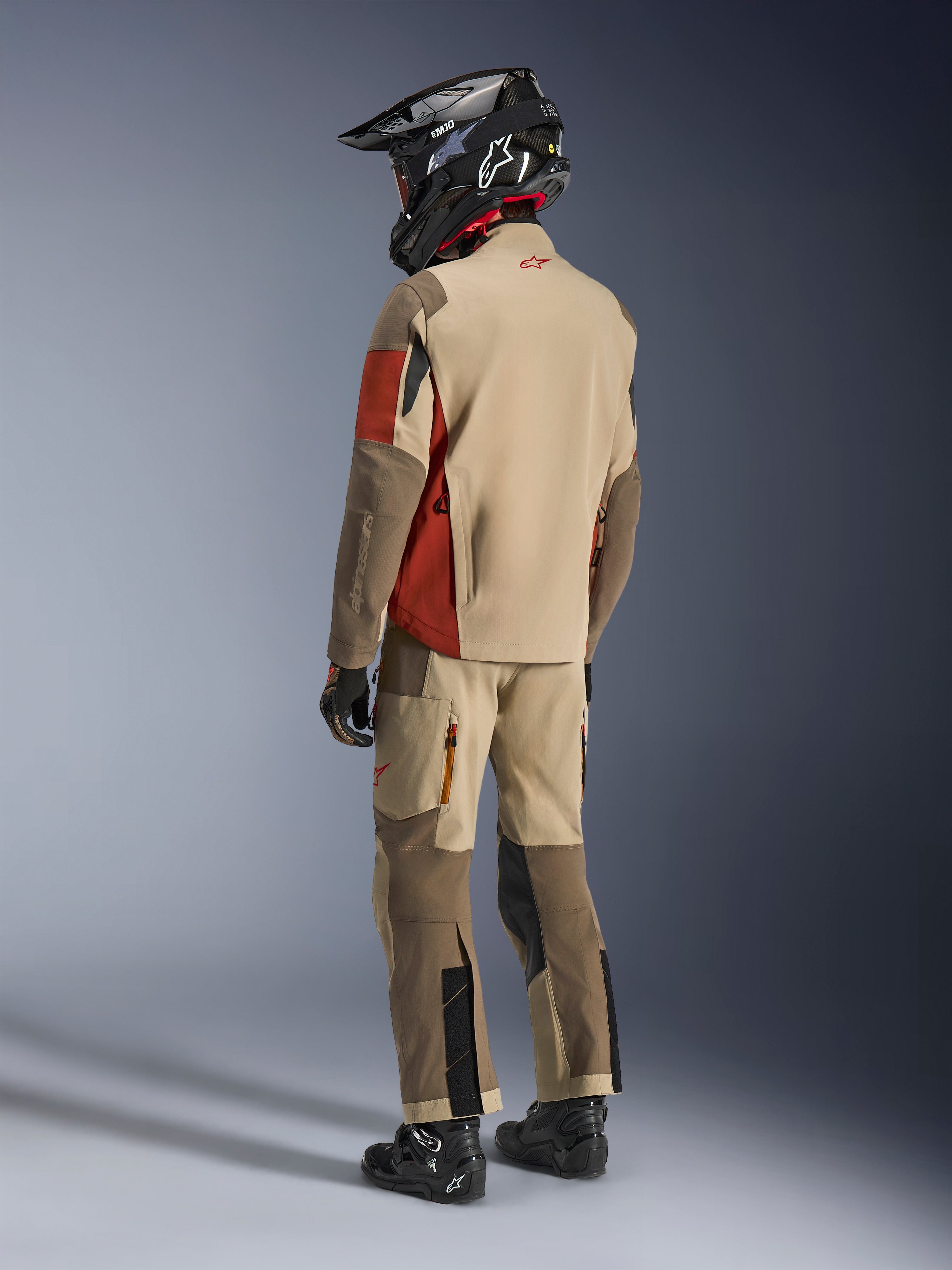 Pilota adventure visto di spalle che indossa la giacca Alpinestars Maxdura Drystar®XF Laminated Armored Shell in Aluminum Walnut Rust Brown, abbinata a pantaloni da moto coordinati, casco off-road nero e stivali neri, outfit tecnico da touring completo.