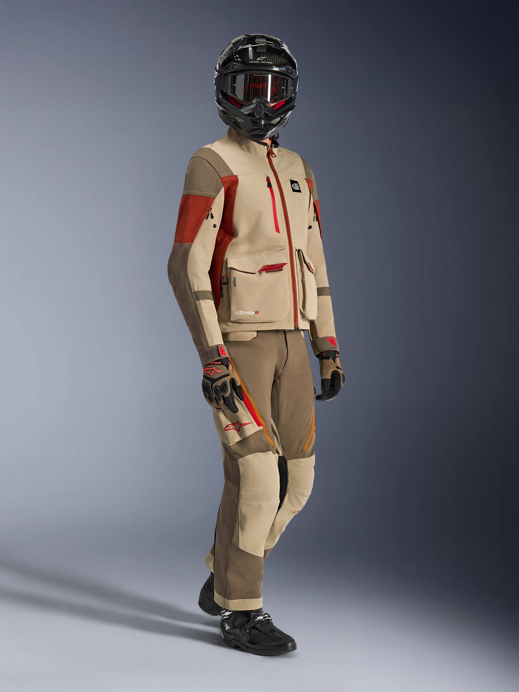 Pilota adventure che indossa la giacca Maxdura Drystar®XF Laminated Armored Shell, colore Aluminum Walnut e Rust Brown, pantaloni coordinati color noce e sabbia, casco in fibra di carbonio nero e stivali da moto neri, abbigliamento tecnico da touring per motociclismo adventure
