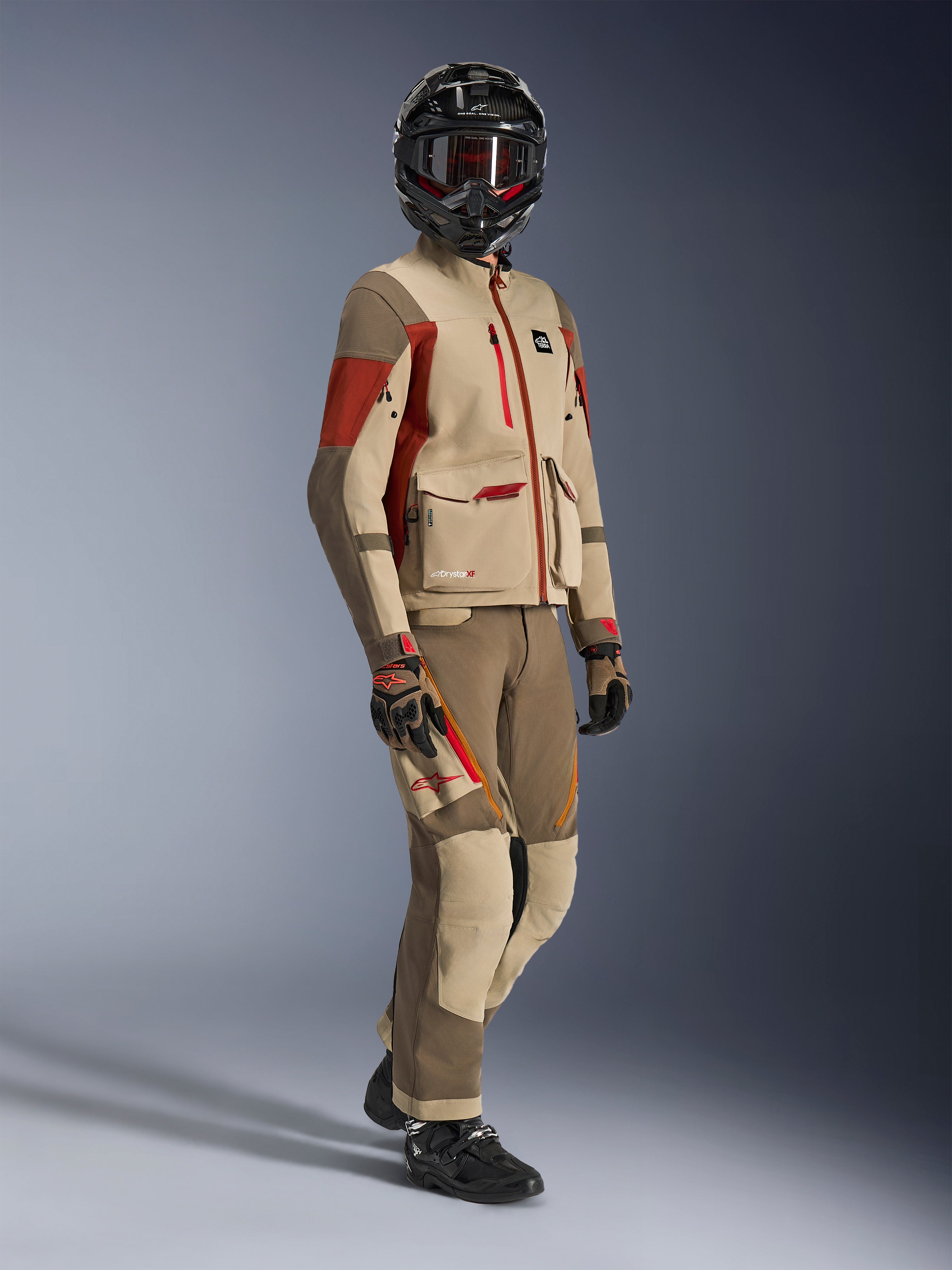Pilota adventure che indossa la giacca Maxdura Drystar®XF Laminated Armored Shell, colore Aluminum Walnut e Rust Brown, pantaloni coordinati color noce e sabbia, casco in fibra di carbonio nero e stivali da moto neri, abbigliamento tecnico da touring per motociclismo adventure