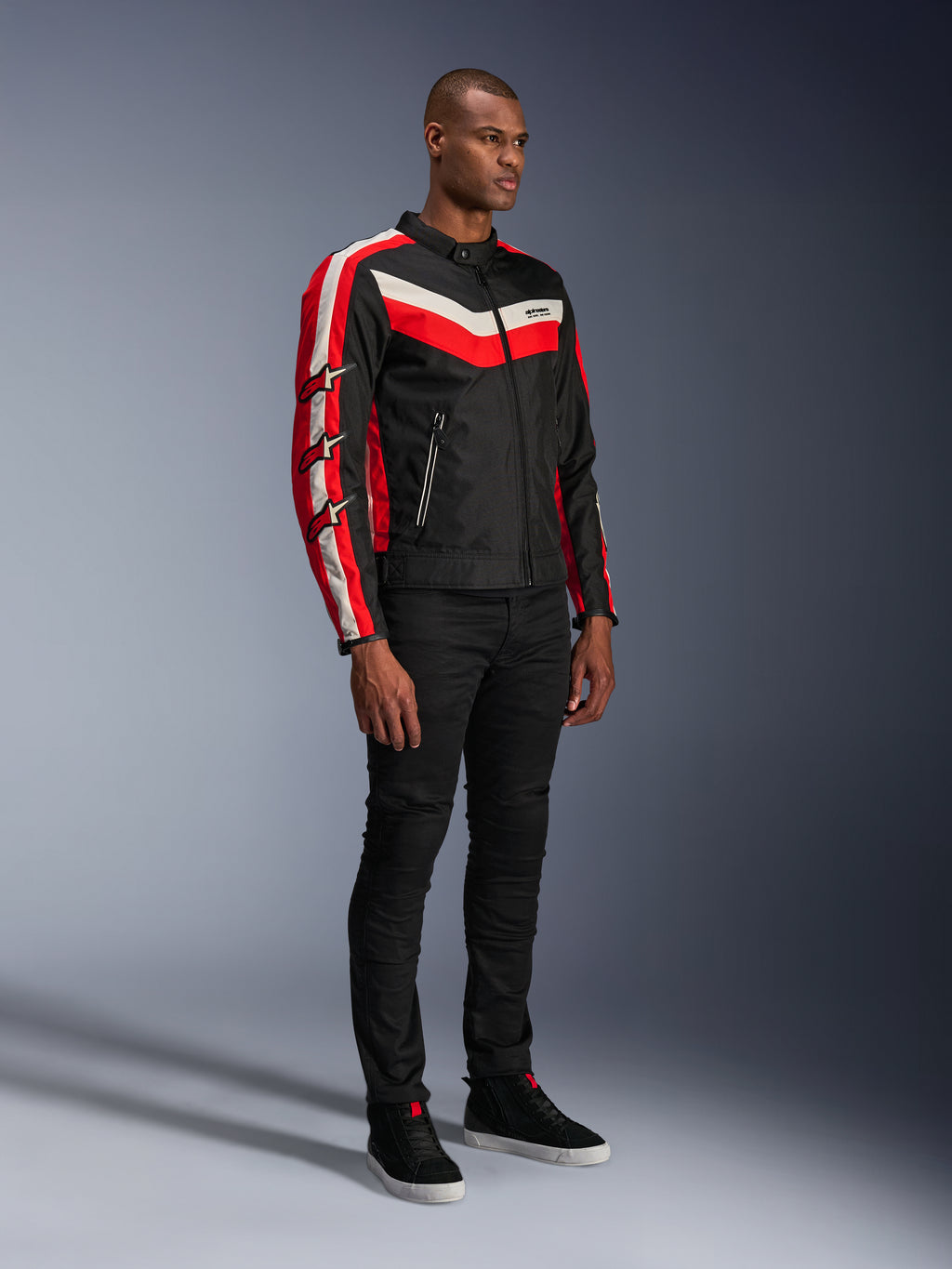 Modello che indossa la giacca Alpinestars T-Dyno WR, giacca da moto in tessuto in Black Bright Red Ecru, con strisce rosse e bianco panna e loghi a stella Alpinestars sulle maniche, abbinata a pantaloni neri e sneakers alte nere, abbigliamento da moto urbano d'ispirazione vintage.