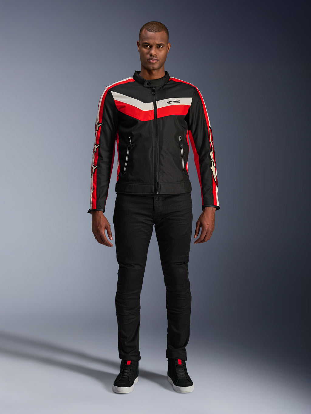 Giacca Alpinestars T-Dyno WR, giacca da moto in tessuto nei colori Nero, Rosso Brillante ed Ecru, con strisce a V sul petto e loghi con stelle sulle maniche, indossata con pantaloni neri e scarpe da guida nere, abbigliamento urban d'ispirazione heritage
