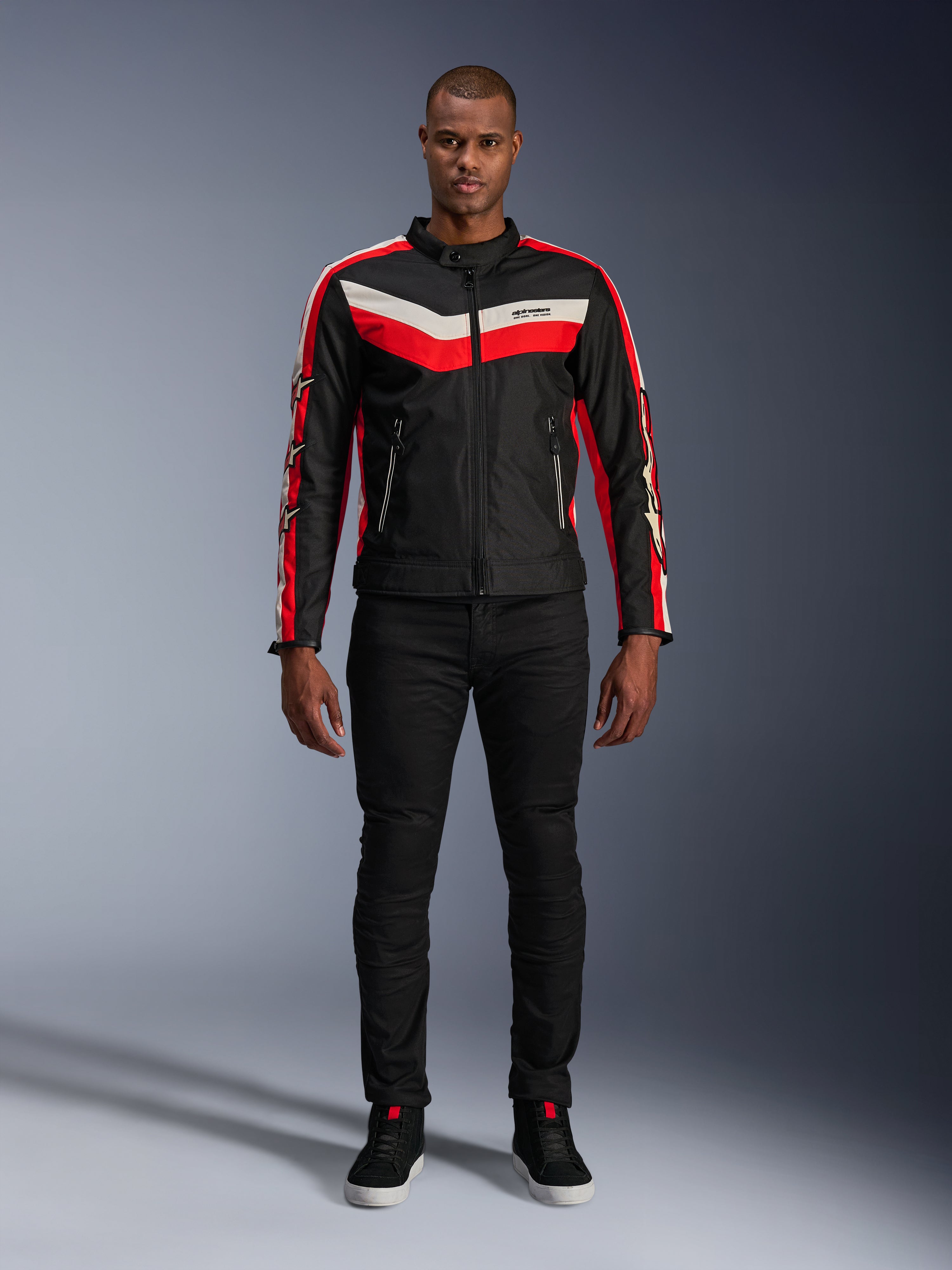 Giacca Alpinestars T-Dyno WR, giacca da moto in tessuto nei colori Nero, Rosso Brillante ed Ecru, con strisce a V sul petto e loghi con stelle sulle maniche, indossata con pantaloni neri e scarpe da guida nere, abbigliamento urban d'ispirazione heritage