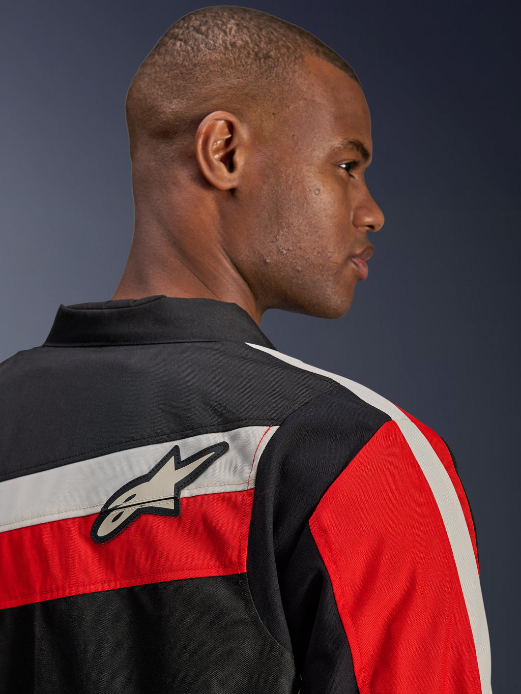 Giacca Alpinestars T-Dyno WR, vista posteriore ravvicinata su un modello, giacca da moto in tessuto nera con strisce orizzontali ecru e rosso acceso, toppa con logo a stella Alpinestars sulla spalla, stile biker d'ispirazione heritage