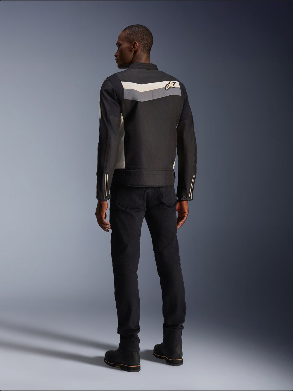 Modello maschile visto da dietro che indossa la giacca Alpinestars T-Dyno WR in Nero Grigio Scuro Ecru, giacca da moto in tessuto con striscia orizzontale e logo a stella sul retro, abbinata a pantaloni da guida e stivali neri, estetica urban motorcycle gear.