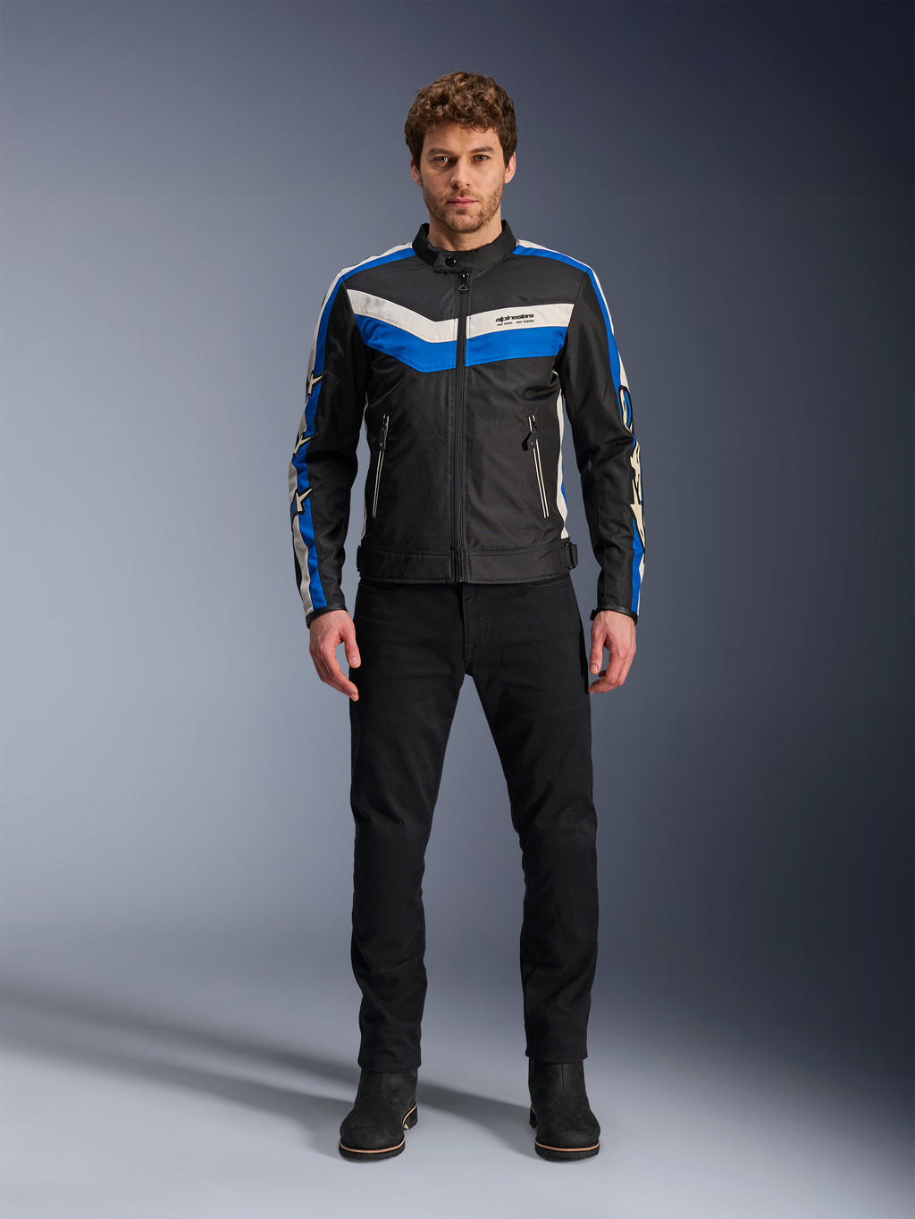 Giacca Alpinestars T-Dyno WR, giacca da moto in tessuto, nero, blu ed ecru, indossata da un modello con pantaloni e stivali neri, con strisce a chevron sul petto e design biker heritage