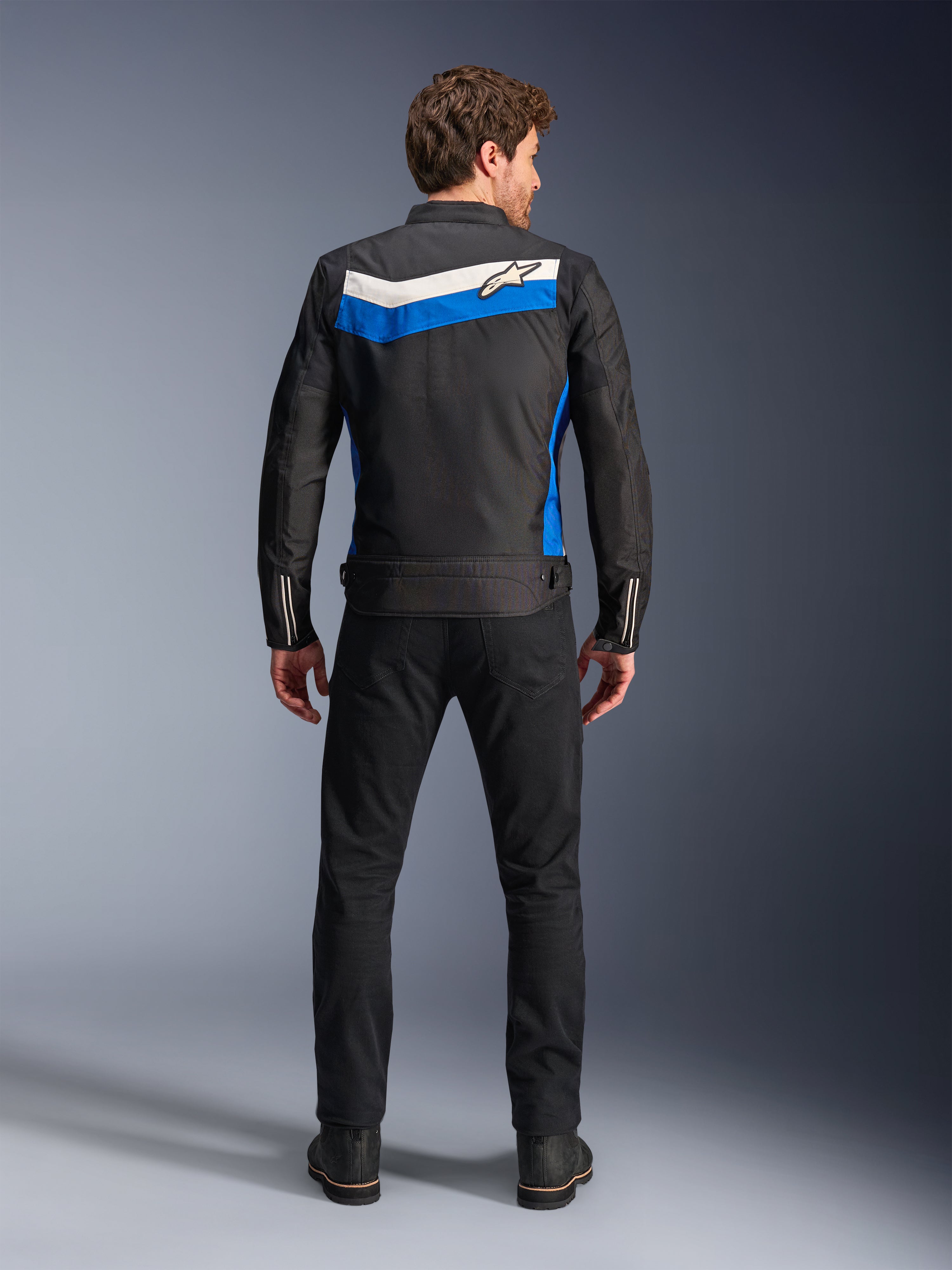 Giacca Alpinestars T-Dyno WR, giacca da moto in tessuto, colorazione Nero Blu Ecru, vista posteriore completa su modello, caratterizzata da una striscia a chevron blu e ecru con logo Alpinestars, abbinata a pantaloni e stivali neri, abbigliamento da moto urbano d'ispirazione vintage