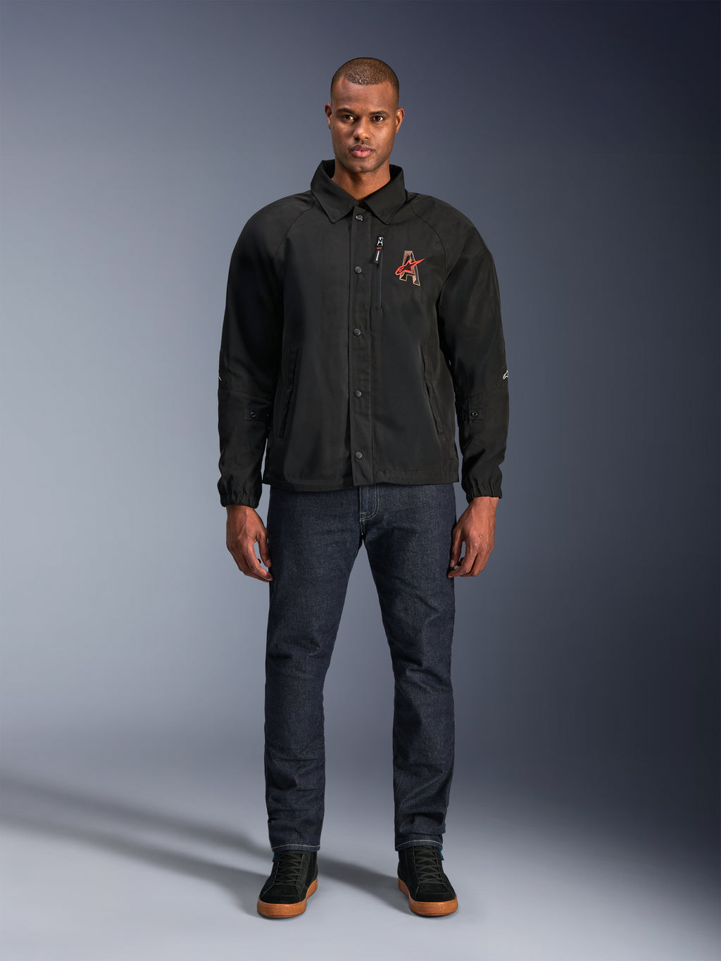 Giacca Alpinestars Revved WR nera, indossata da un modello con jeans blu scuro e sneakers nere, vista frontale, con chiusura a bottoni a pressione e logo ricamato in 3D sul petto, abbigliamento da moto urban contemporary