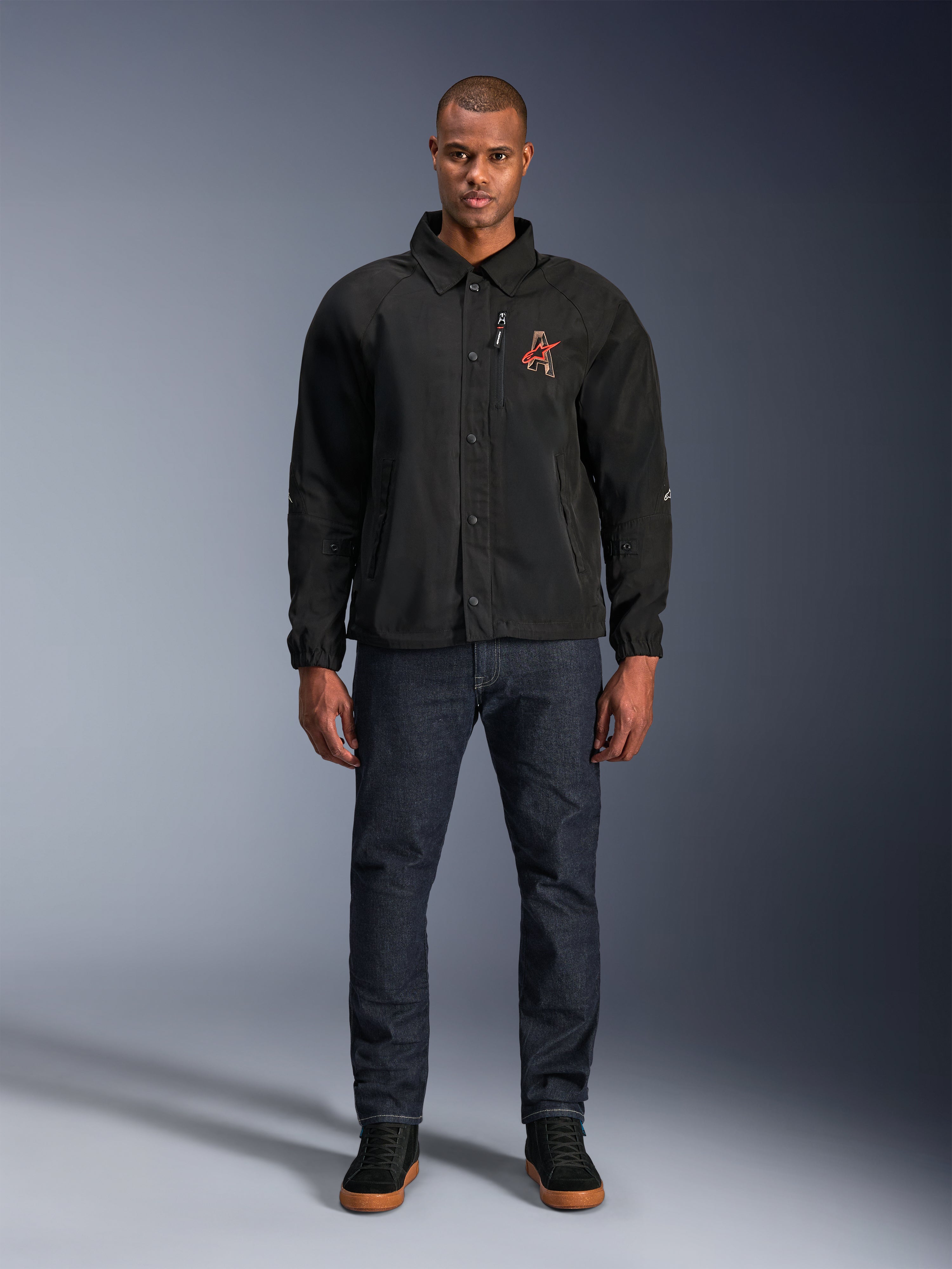 Giacca Alpinestars Revved WR nera, indossata da un modello con jeans blu scuro e sneakers nere, vista frontale, con chiusura a bottoni a pressione e logo ricamato in 3D sul petto, abbigliamento da moto urban contemporary