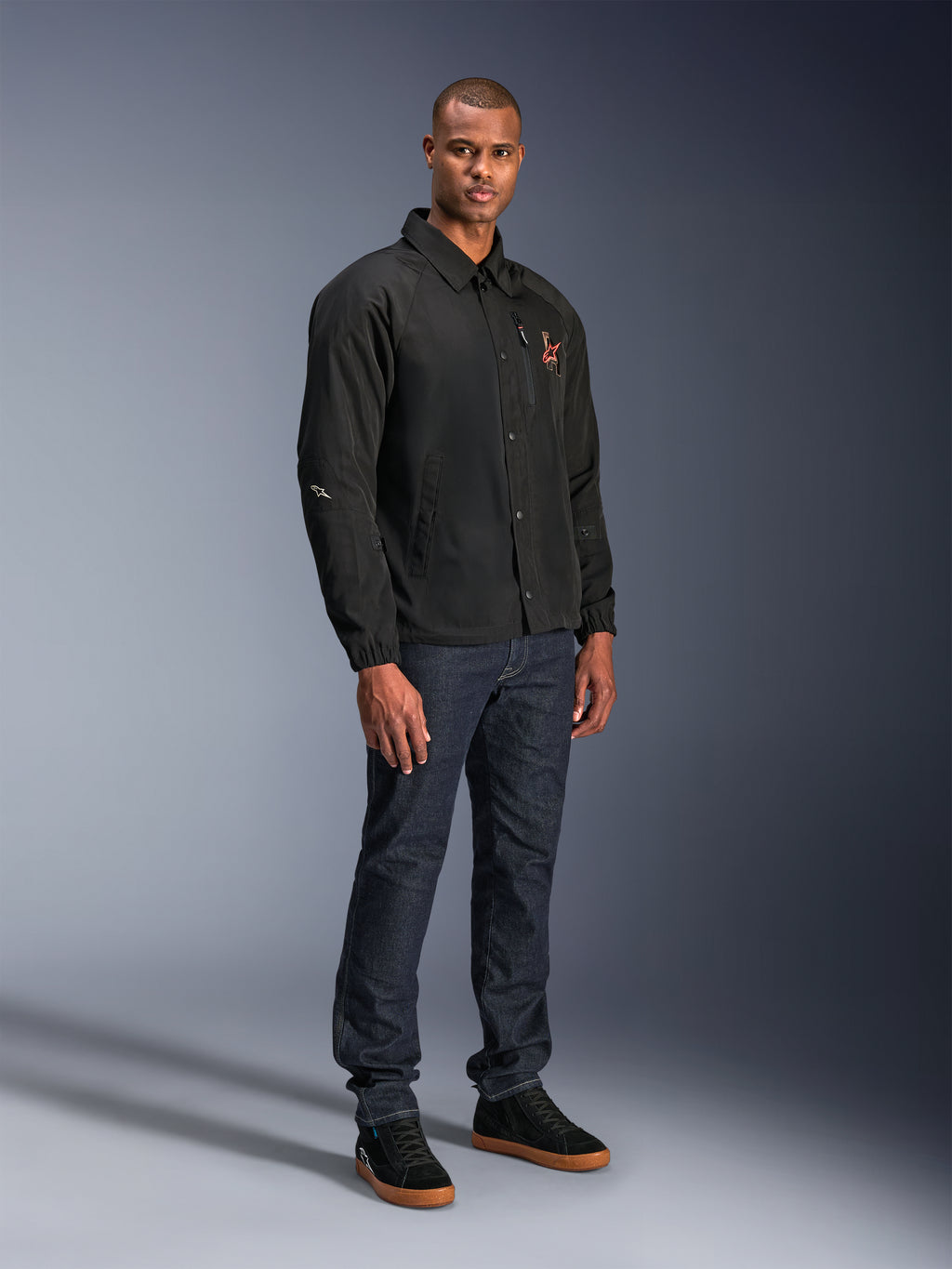 Giacca Alpinestars Revved WR in nero, giacca da moto in stile coach urbano indossata da un modello con jeans blu scuro e sneakers nere, con chiusura frontale a bottoni a pressione e logo ricamato, design minimalista e sportivo per la guida su strada