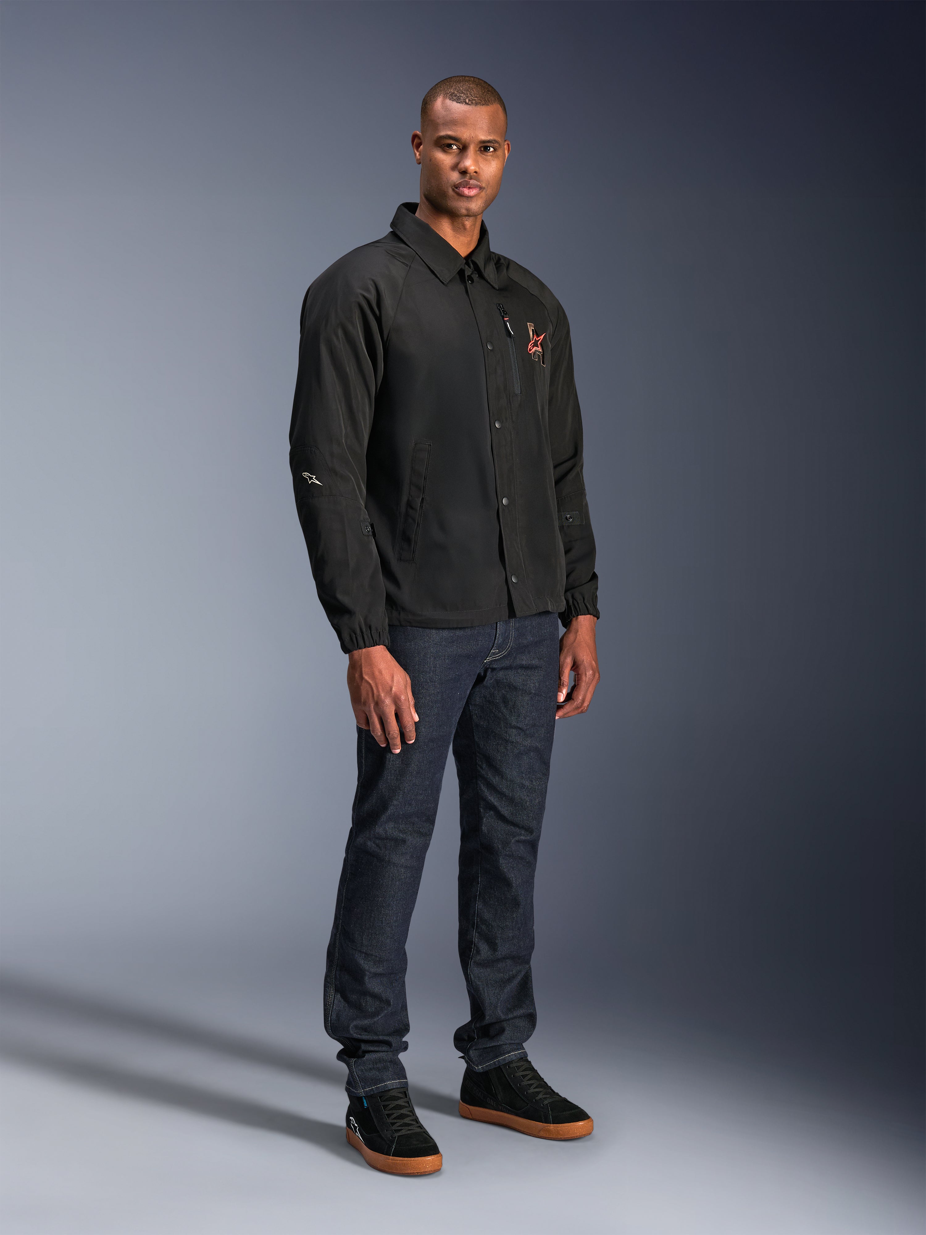Giacca Alpinestars Revved WR in nero, giacca da moto in stile coach urbano indossata da un modello con jeans blu scuro e sneakers nere, con chiusura frontale a bottoni a pressione e logo ricamato, design minimalista e sportivo per la guida su strada