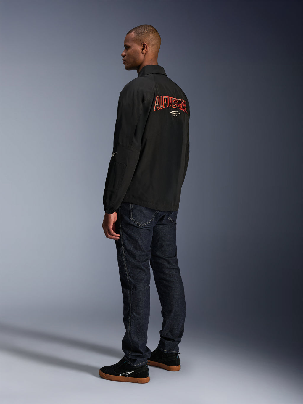 Giacca Alpinestars Revved WR, nero, con grande logo Alpinestars arancione ricamato in 3D sul retro, vista posteriore a tre quarti di un modello che indossa jeans blu scuro e sneakers nere, giacca da moto urban contemporary con silhouette stile giacca da allenatore