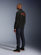 Giacca Alpinestars Revved WR, nero, con grande logo Alpinestars arancione ricamato in 3D sul retro, vista posteriore a tre quarti di un modello che indossa jeans blu scuro e sneakers nere, giacca da moto urban contemporary con silhouette stile giacca da allenatore