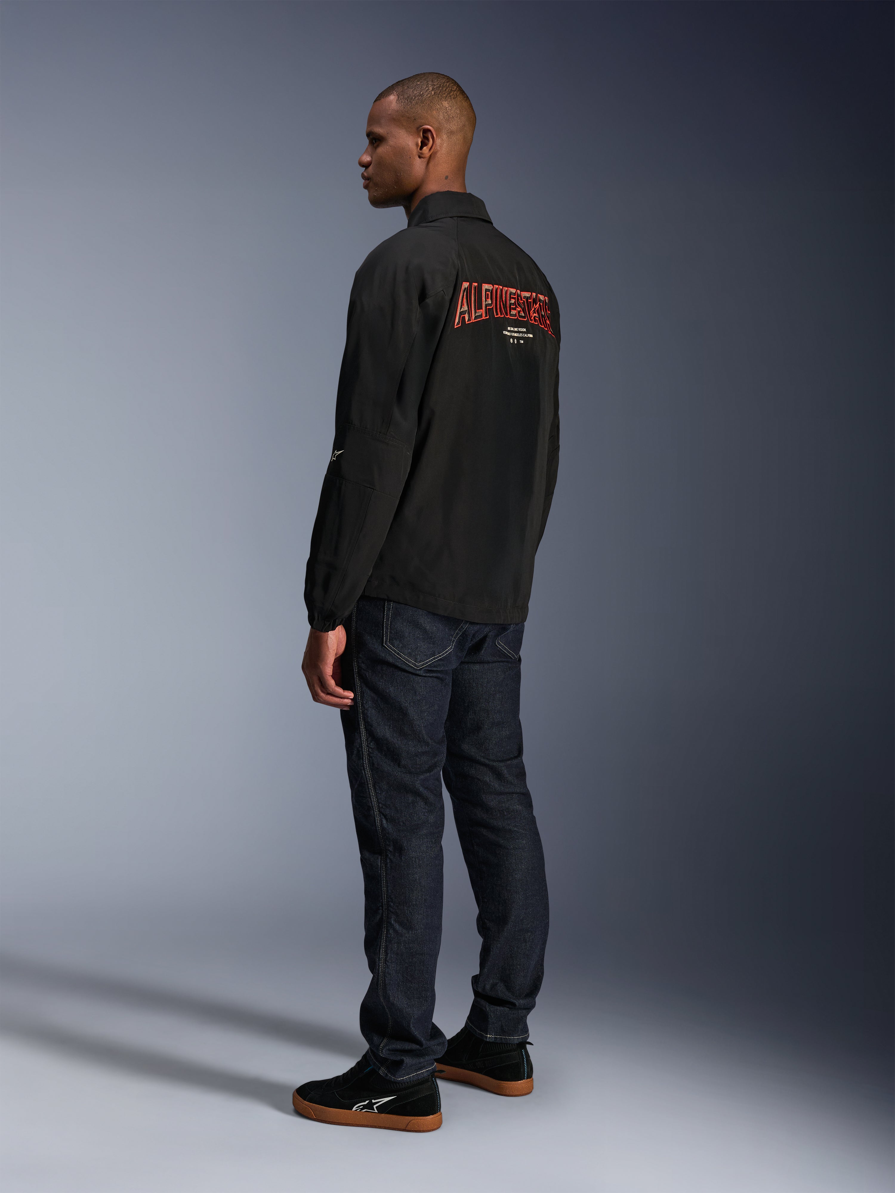 Giacca Alpinestars Revved WR, nero, con grande logo Alpinestars arancione ricamato in 3D sul retro, vista posteriore a tre quarti di un modello che indossa jeans blu scuro e sneakers nere, giacca da moto urban contemporary con silhouette stile giacca da allenatore