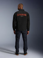 Alpinestars Giacca Revved WR, nera, vista posteriore con grande ricamo rosso 'ALPINESTARS' effetto 3D, giacca stile urban coach, indossata con jeans denim scuro e sneakers nere, abbigliamento da moto minimalista