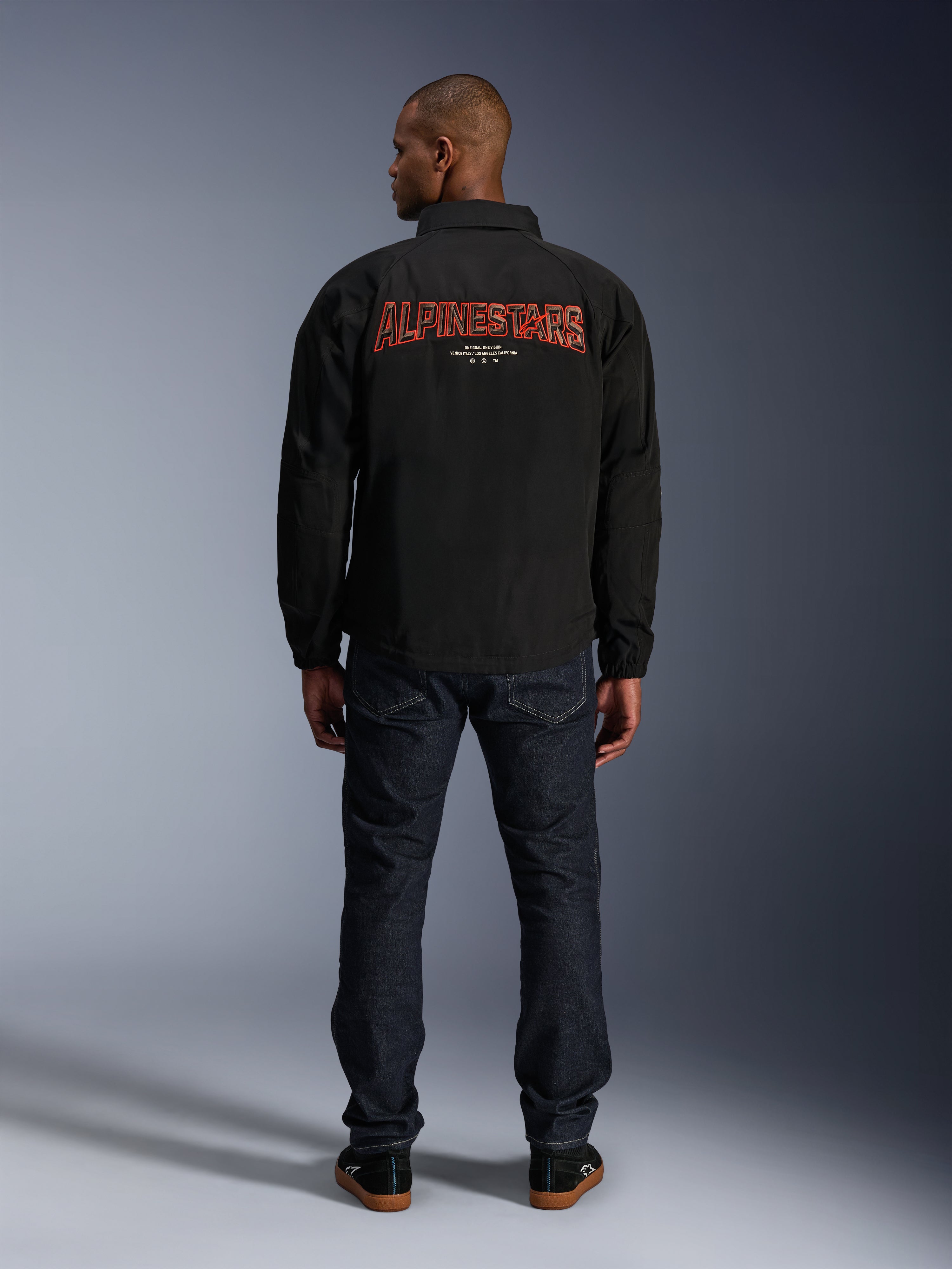 Alpinestars Giacca Revved WR, nera, vista posteriore con grande ricamo rosso 'ALPINESTARS' effetto 3D, giacca stile urban coach, indossata con jeans denim scuro e sneakers nere, abbigliamento da moto minimalista
