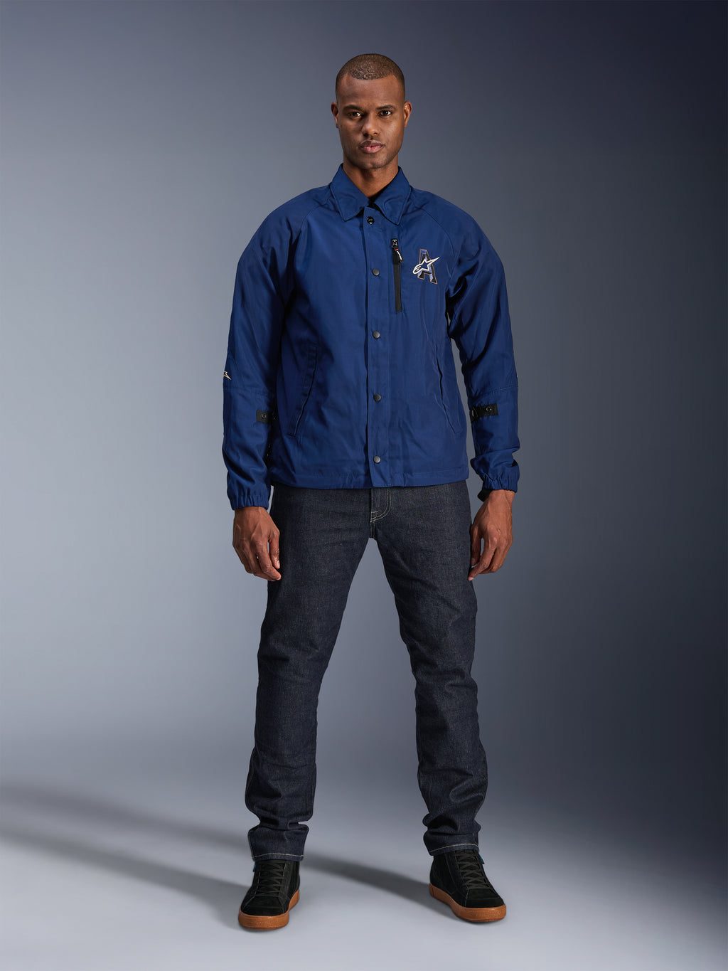 Giacca Alpinestars Revved WR, blu navy, giacca da moto stile coach, indossata da un uomo con jeans in denim scuro e sneakers nere, con chiusura a bottoni a pressione e logo Alpinestars 3D, streetwear contemporaneo urbano