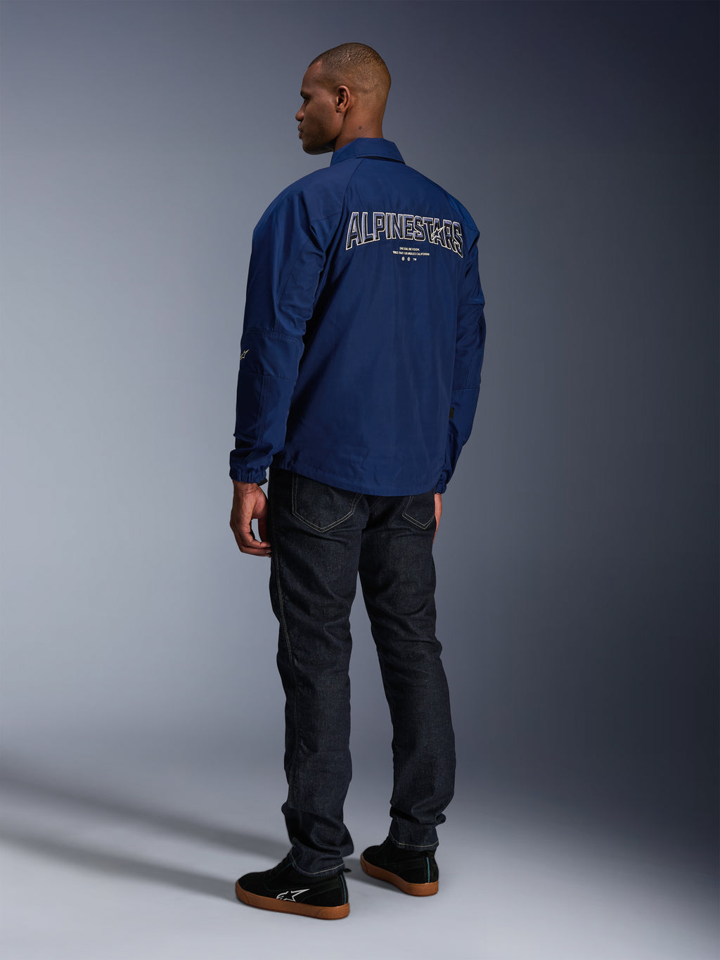 Modello che indossa la giacca Alpinestars Revved WR in blu navy vista da dietro, con grande ricamo 'ALPINESTARS' profilato in bianco sulla schiena, abbinata a pantaloni in denim scuro e sneaker nere, estetica motociclistica urbana.