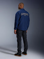 Modello che indossa la giacca Alpinestars Revved WR in blu navy vista da dietro, con grande ricamo 'ALPINESTARS' profilato in bianco sulla schiena, abbinata a pantaloni in denim scuro e sneaker nere, estetica motociclistica urbana.