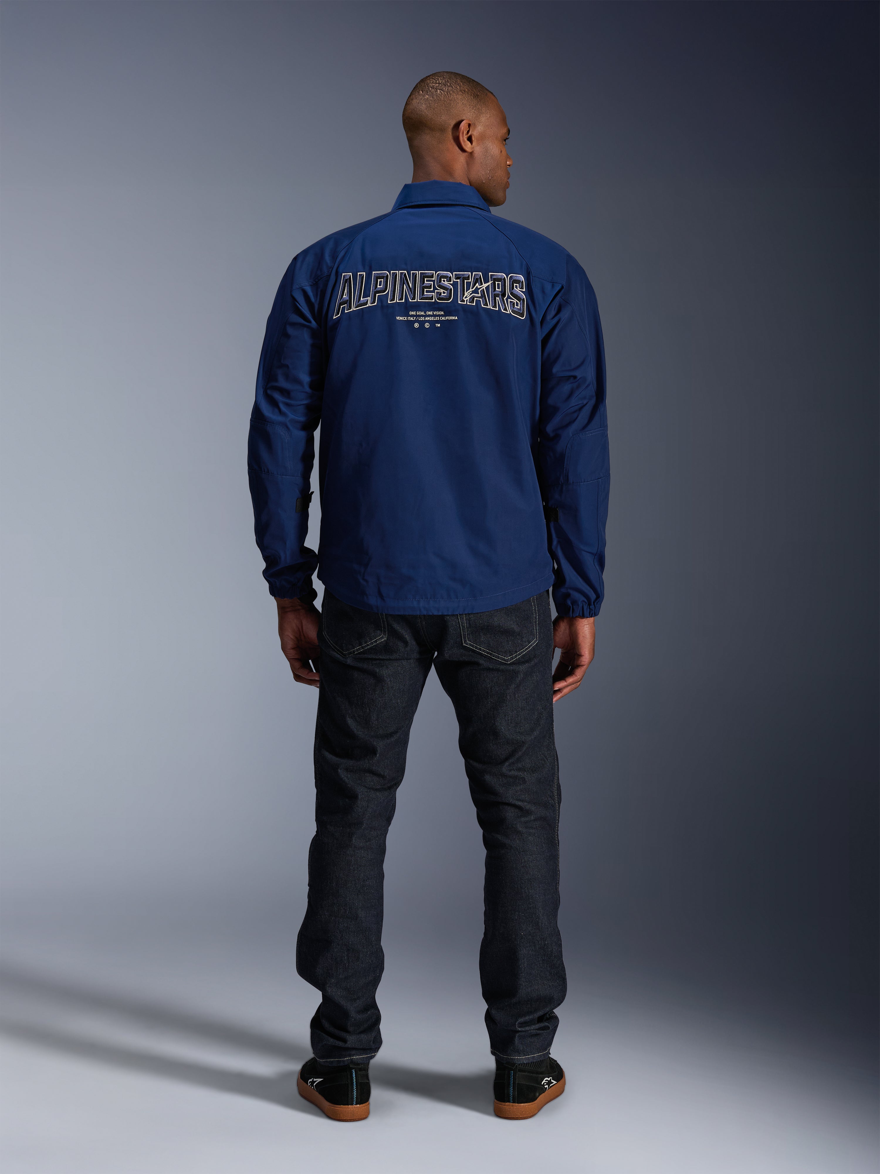 Giacca Alpinestars Revved WR in blu navy, vista posteriore, con grande ricamo 'ALPINESTARS' effetto 3D sulla schiena, abbinata a jeans in denim scuro e sneakers nere, abbigliamento moto urban.