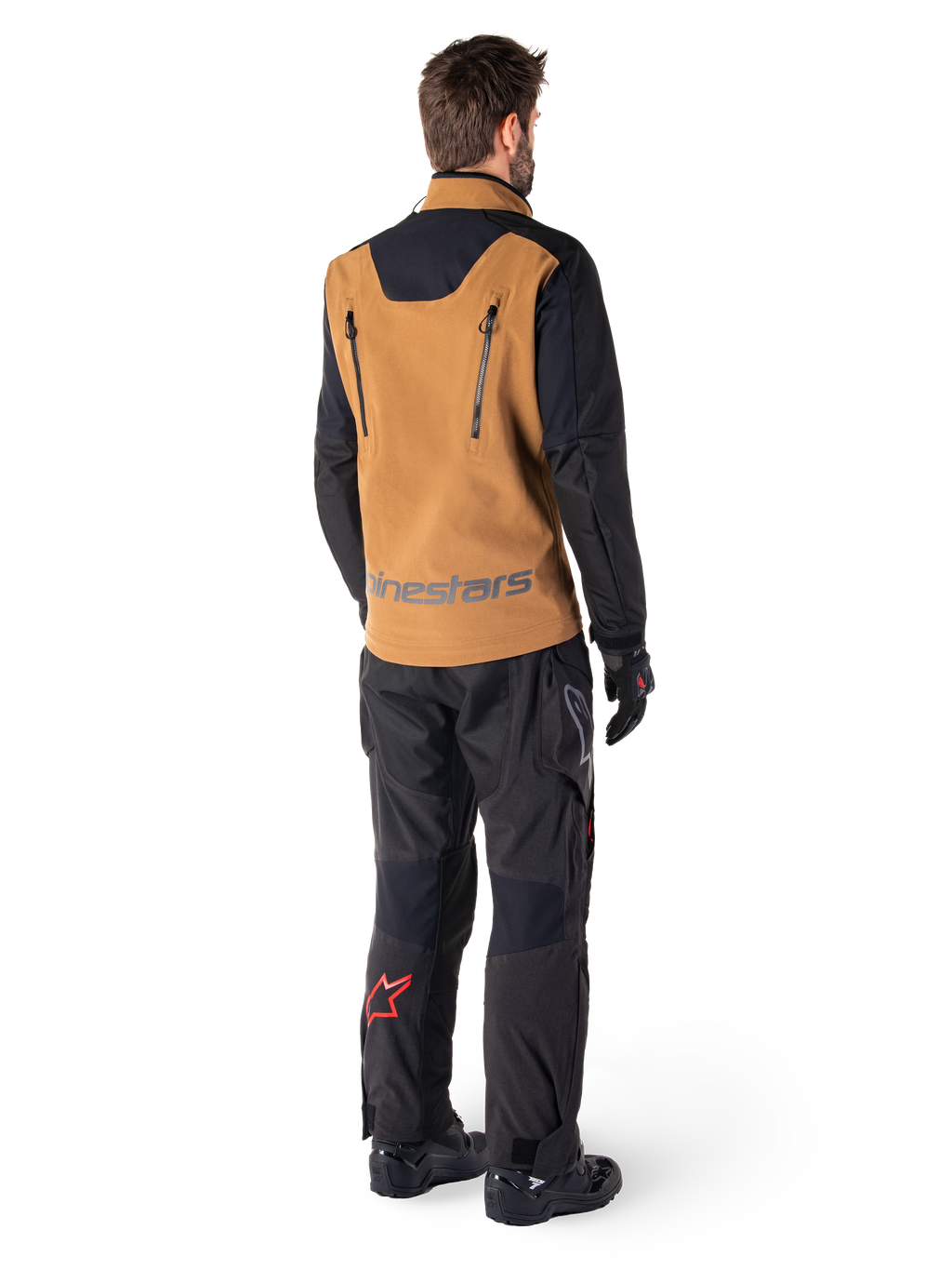 Giacca Alpinestars Hyde XT Stretch Drystar®XF e pantaloni Hyde XT Drystar®, vista posteriore di un pilota che indossa abbigliamento adventure touring tan e nero, giacca con cerniere verticali sulla schiena e marchio Alpinestars, pantaloni neri con logo a stella rosso sul polpaccio, abbinati a guanti e stivali neri.