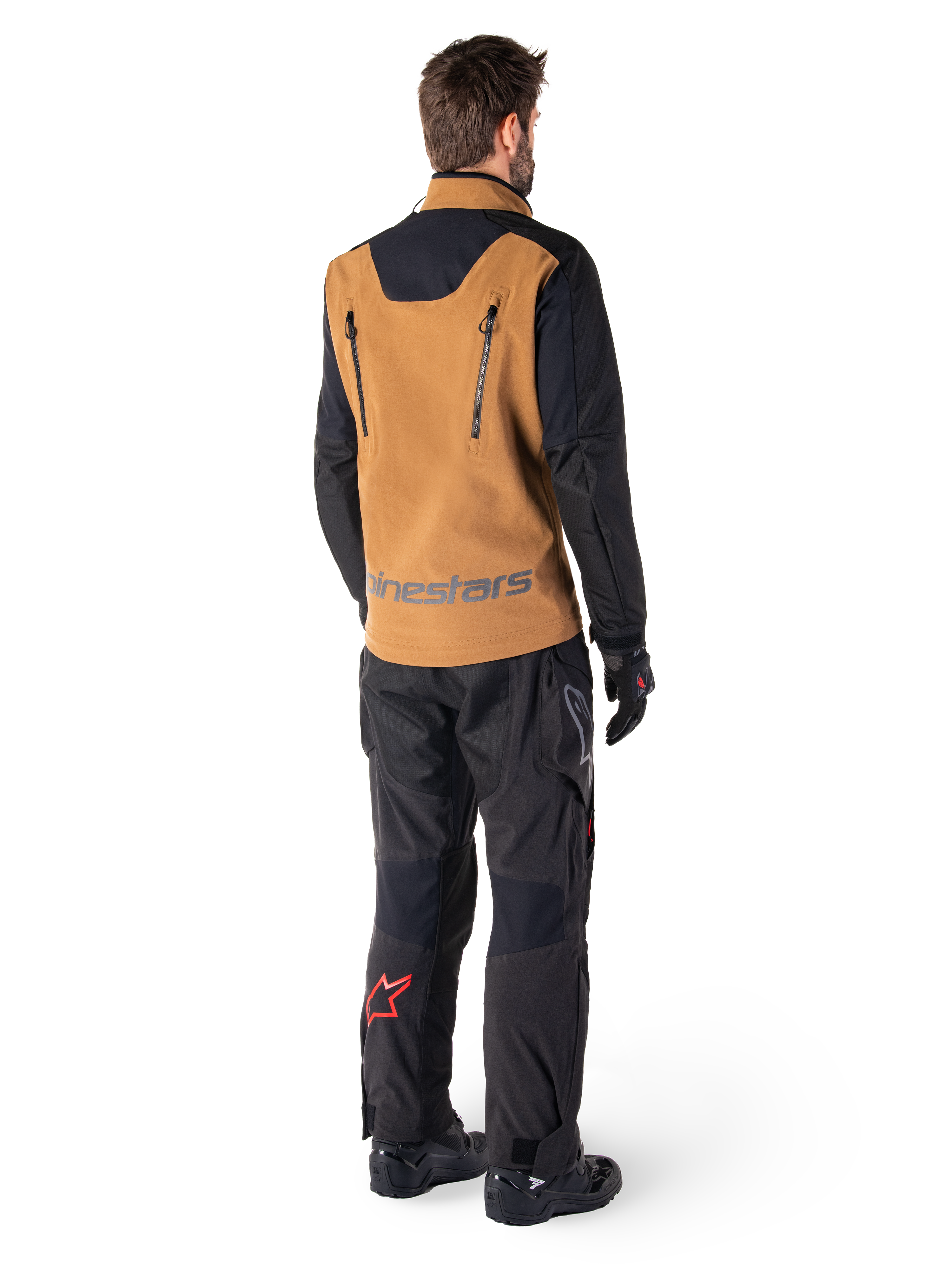 Giacca Alpinestars Hyde XT Stretch Drystar®XF e pantaloni Hyde XT Drystar®, vista posteriore di un pilota che indossa abbigliamento adventure touring tan e nero, giacca con cerniere verticali sulla schiena e marchio Alpinestars, pantaloni neri con logo a stella rosso sul polpaccio, abbinati a guanti e stivali neri.