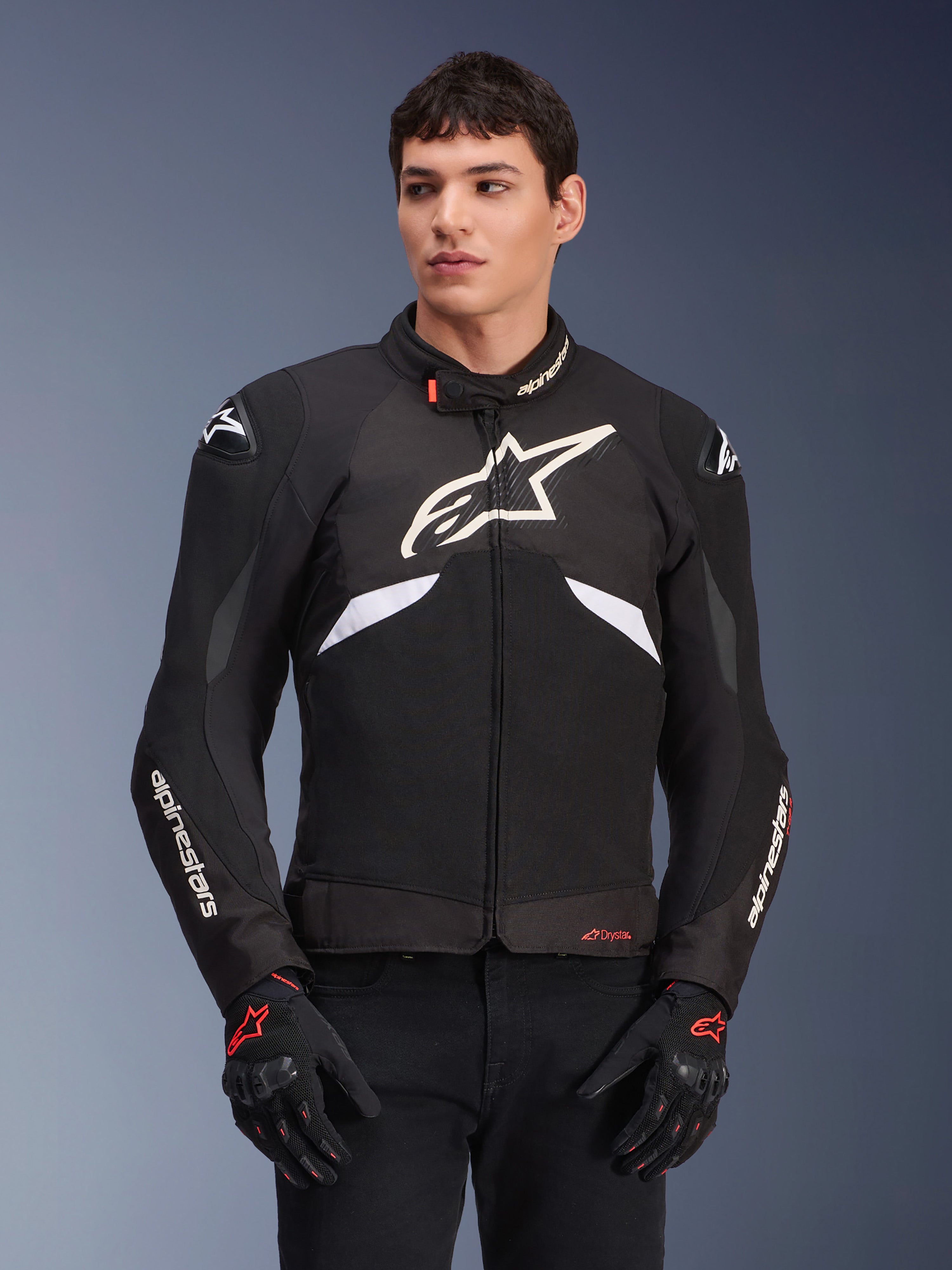 Giacca Alpinestars T-GP R V3 Drystar, giacca sportiva in tessuto impermeabile, nero e bianco con accenti rossi, con un grande logo a stella bianco sul petto e branding bianco sulle maniche, indossata con guanti da moto neri e rossi