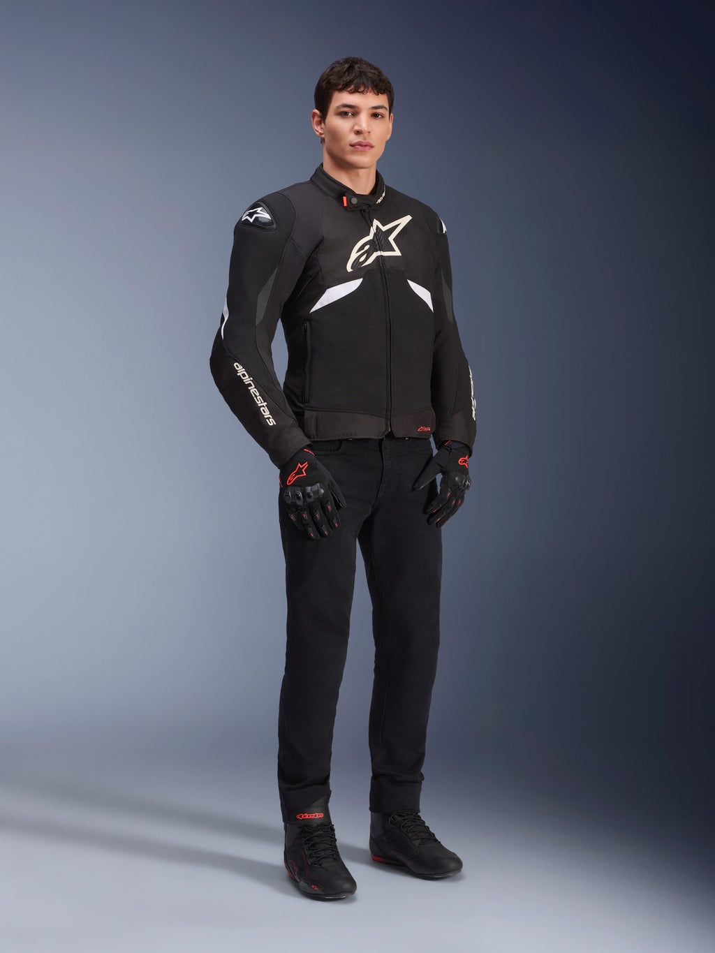 Alpinestars T-GP R V3 Drystar® Jacket in nero e bianco, indossata da un modello con guanti e pantaloni da moto neri, con logo bianco sul petto, dettagli bianchi sulle maniche e slider protettivi sulle spalle, giacca sportiva in tessuto impermeabile per la guida su strada