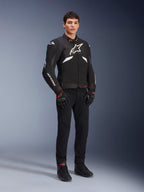 Alpinestars T-GP R V3 Drystar® Jacket in nero e bianco, indossata da un modello con guanti e pantaloni da moto neri, con logo bianco sul petto, dettagli bianchi sulle maniche e slider protettivi sulle spalle, giacca sportiva in tessuto impermeabile per la guida su strada