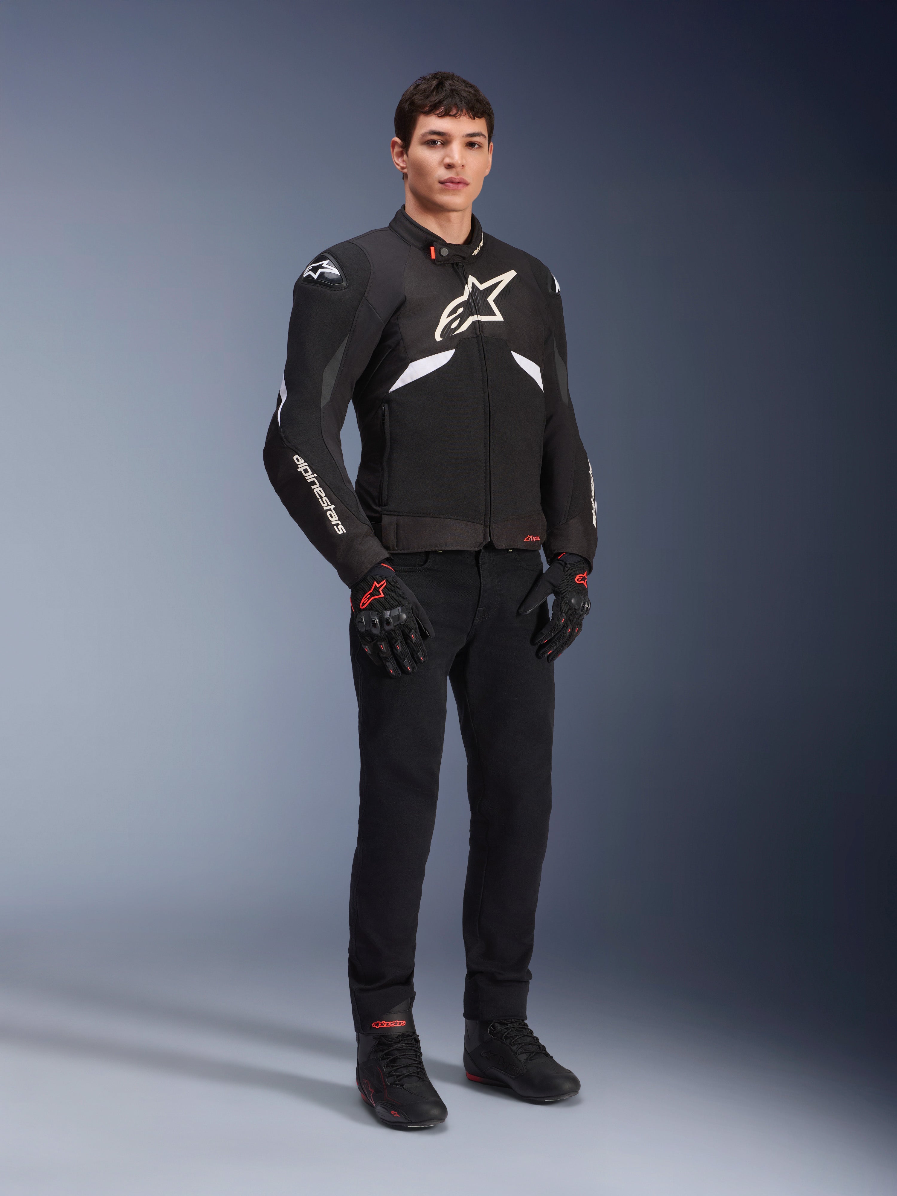 Alpinestars T-GP R V3 Drystar® Jacket in nero e bianco, indossata da un modello con guanti e pantaloni da moto neri, con logo bianco sul petto, dettagli bianchi sulle maniche e slider protettivi sulle spalle, giacca sportiva in tessuto impermeabile per la guida su strada