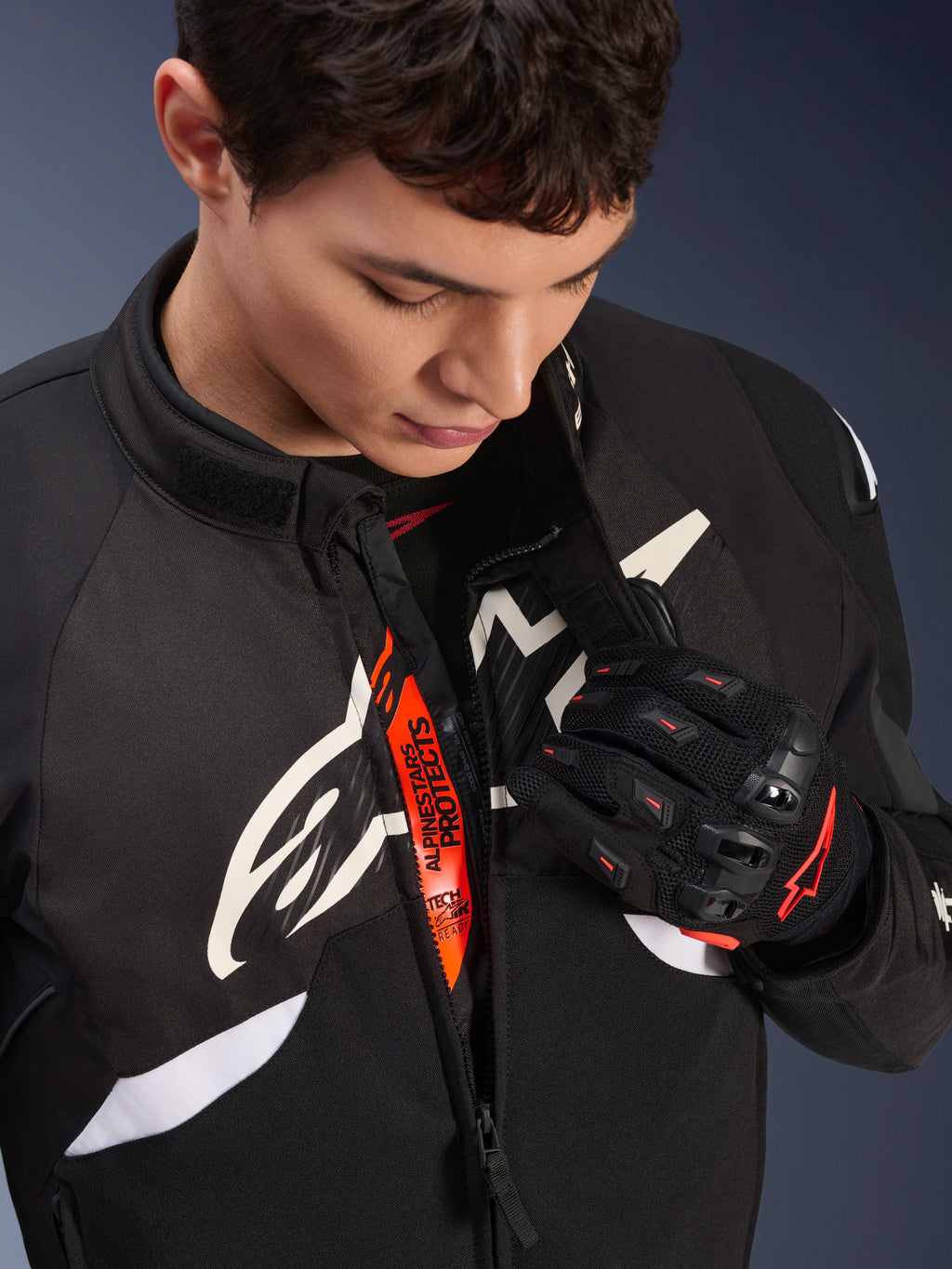 Giacca Alpinestars T-GP R V3 Drystar®, giacca sportiva da moto in tessuto nero e bianco, parzialmente aperta per rivelare la fascia interna arancione 'Alpinestars Protects', indossata con guanti in mesh neri e rossi, abbigliamento protettivo di derivazione racing.