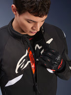 Giacca Alpinestars T-GP R V3 Drystar®, giacca sportiva da moto in tessuto nero e bianco, parzialmente aperta per rivelare la fascia interna arancione 'Alpinestars Protects', indossata con guanti in mesh neri e rossi, abbigliamento protettivo di derivazione racing.